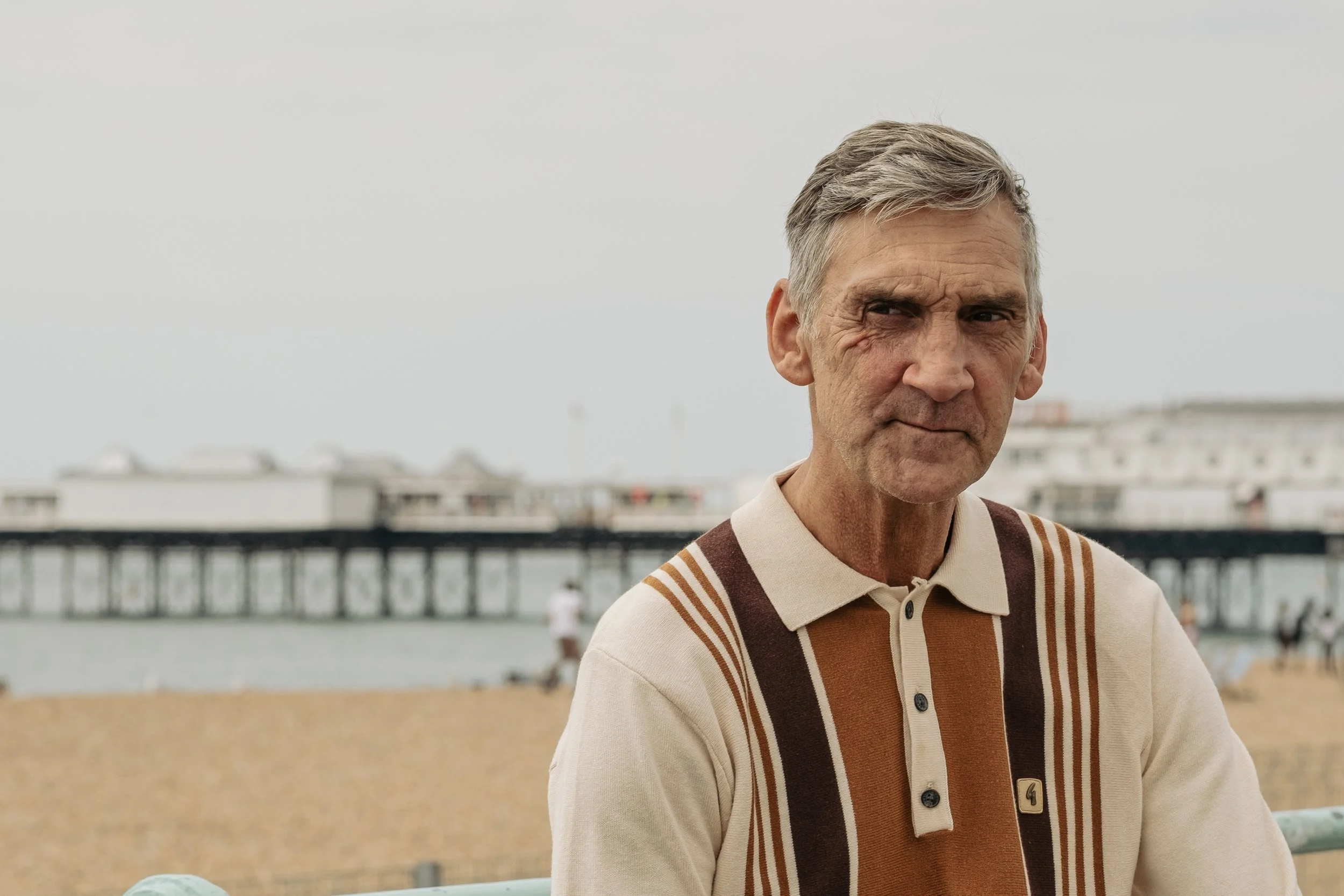 Older man with gray hair and a wrinkled face wearing a beige, brown, and orange striped polo shirt, standing outdoors near a beach with a pier and boats in the background.