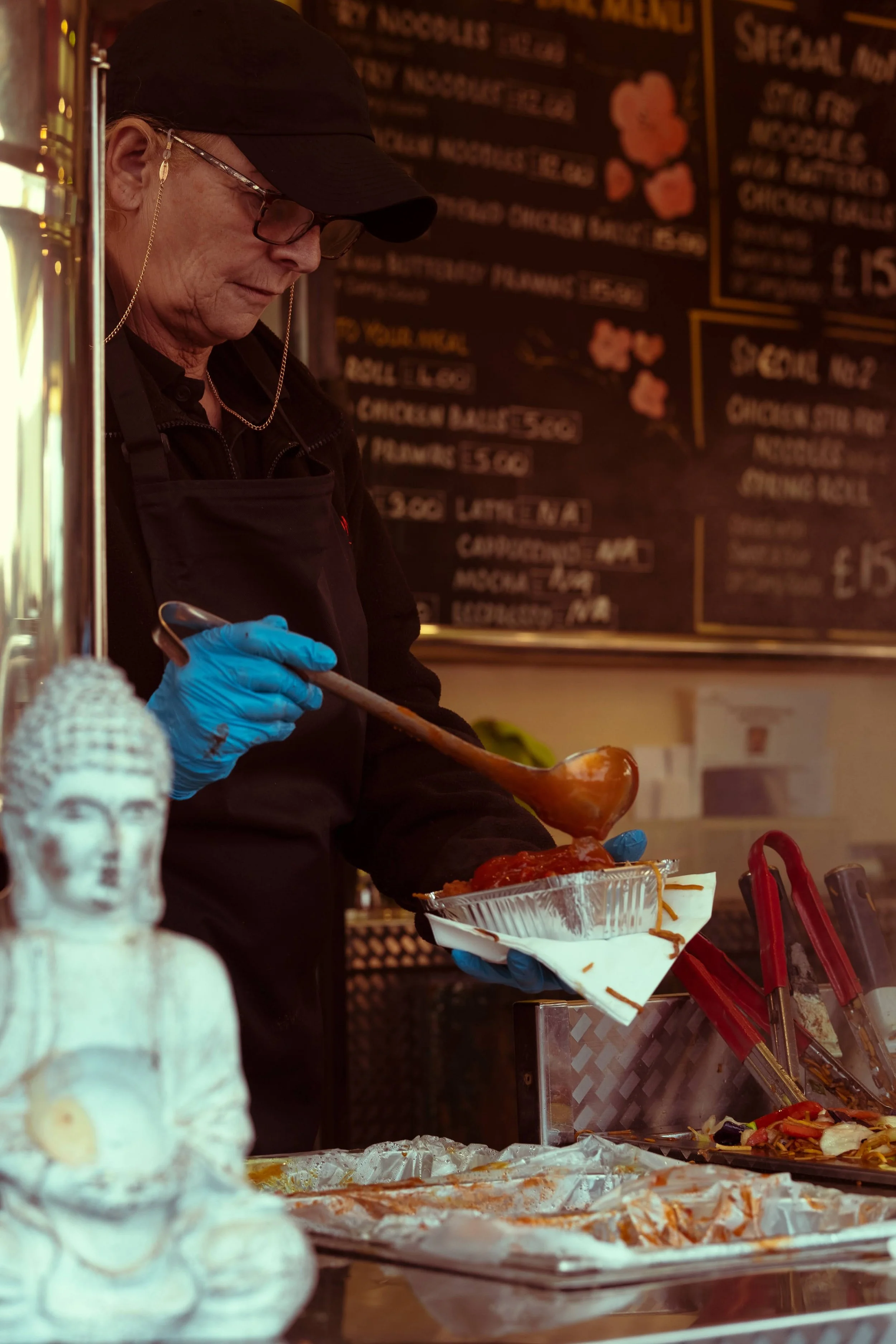A person wearing a black cap, glasses, and blue gloves serving food from a metal tray at a food stand or restaurant. In the foreground, there is a Buddha statue. A menu with food items can be seen in the background.
