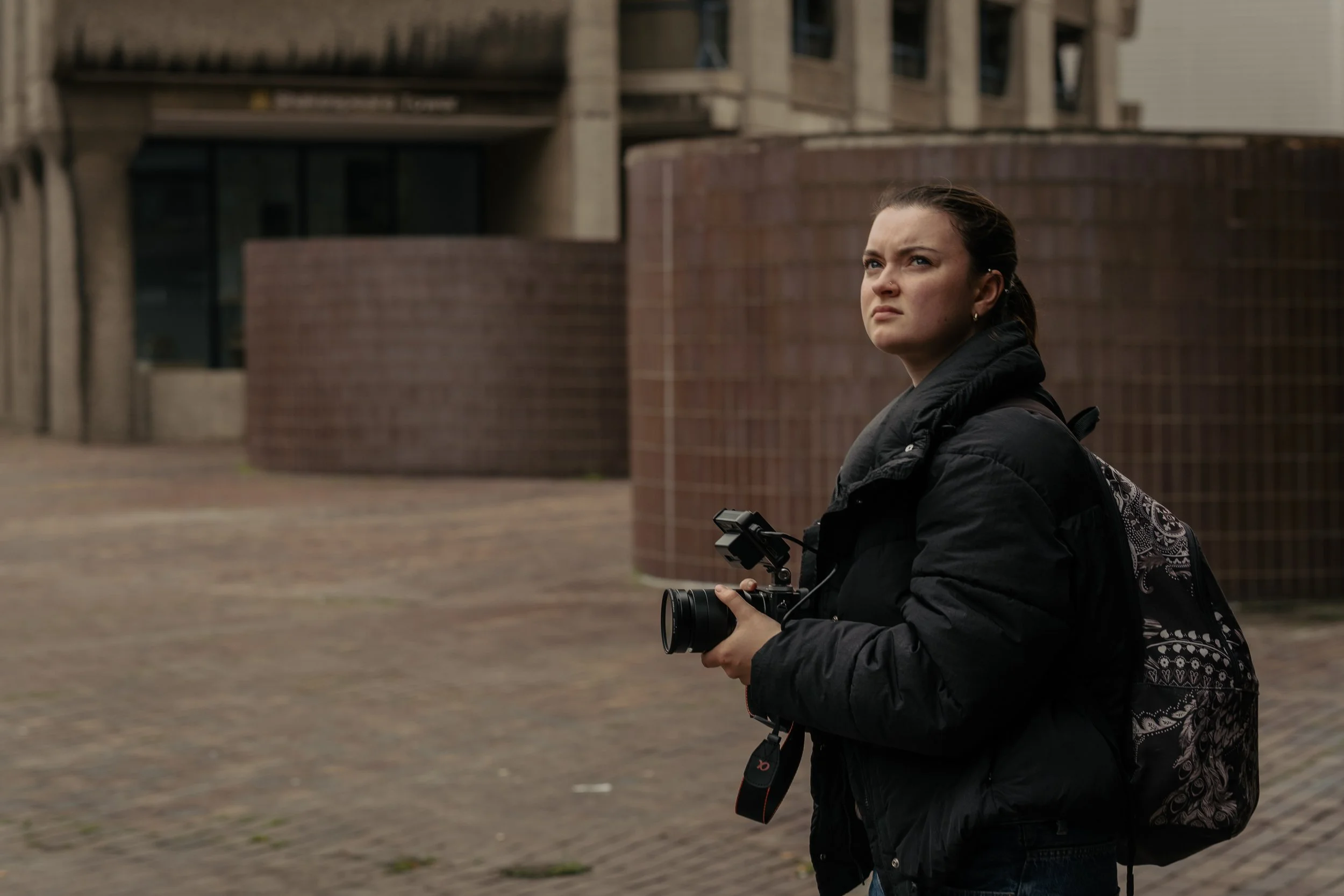 A young woman with a camera in hand stands outdoors in an urban area, wearing a black jacket and backpack, looking contemplative.