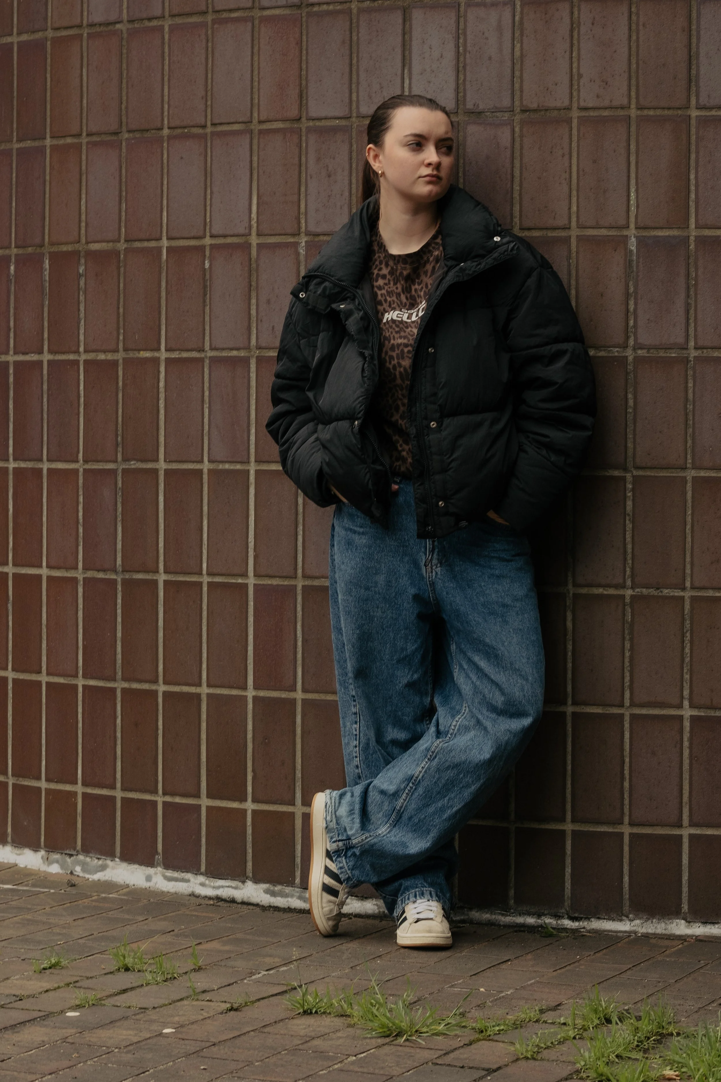 A young woman leaning against a brick wall, wearing a black puffer jacket, leopard print shirt, loose blue jeans, and Adidas sneakers. She has her hands in her pockets and looks to the side.