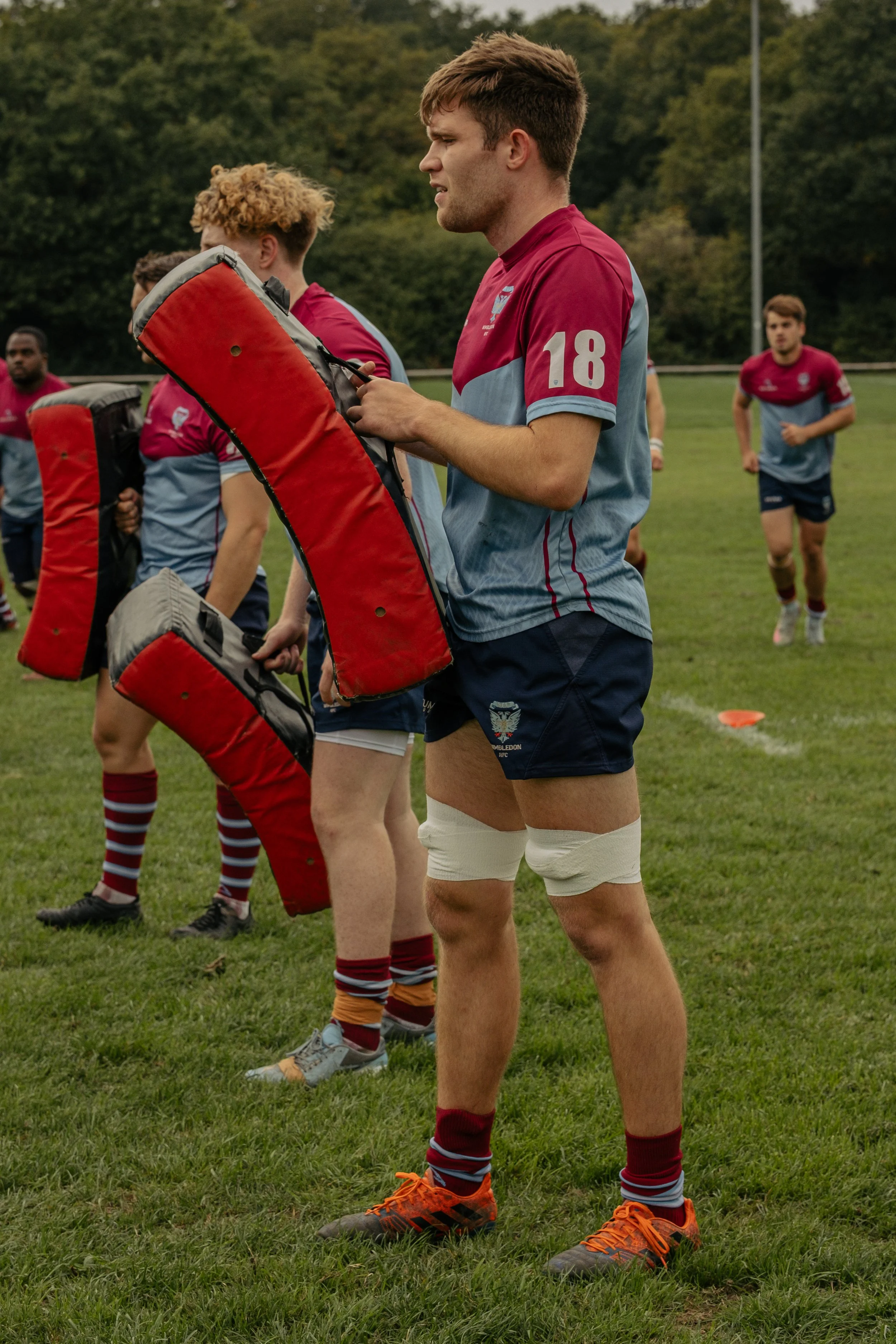 Rugby players preparing for practice or a game on a grassy field, with a player adjusting his gear near other players in the background.