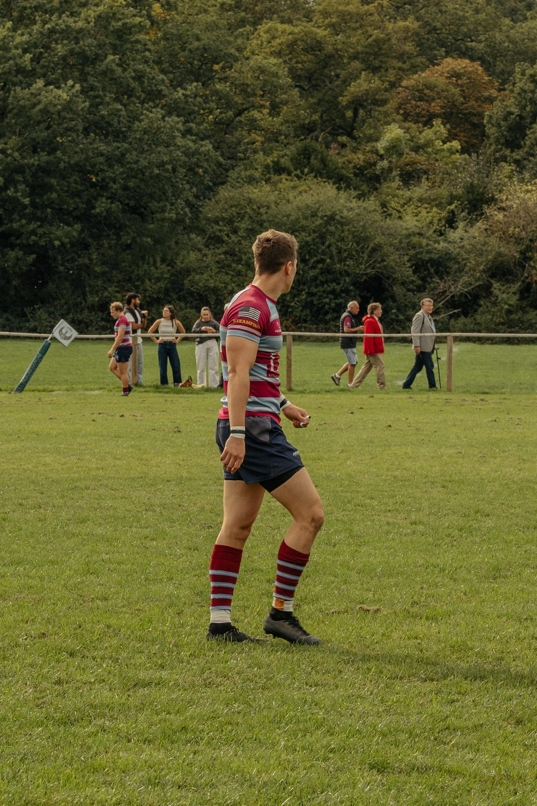 A rugby player in a maroon and gray uniform jogging on a grassy field with spectators and trees in the background.