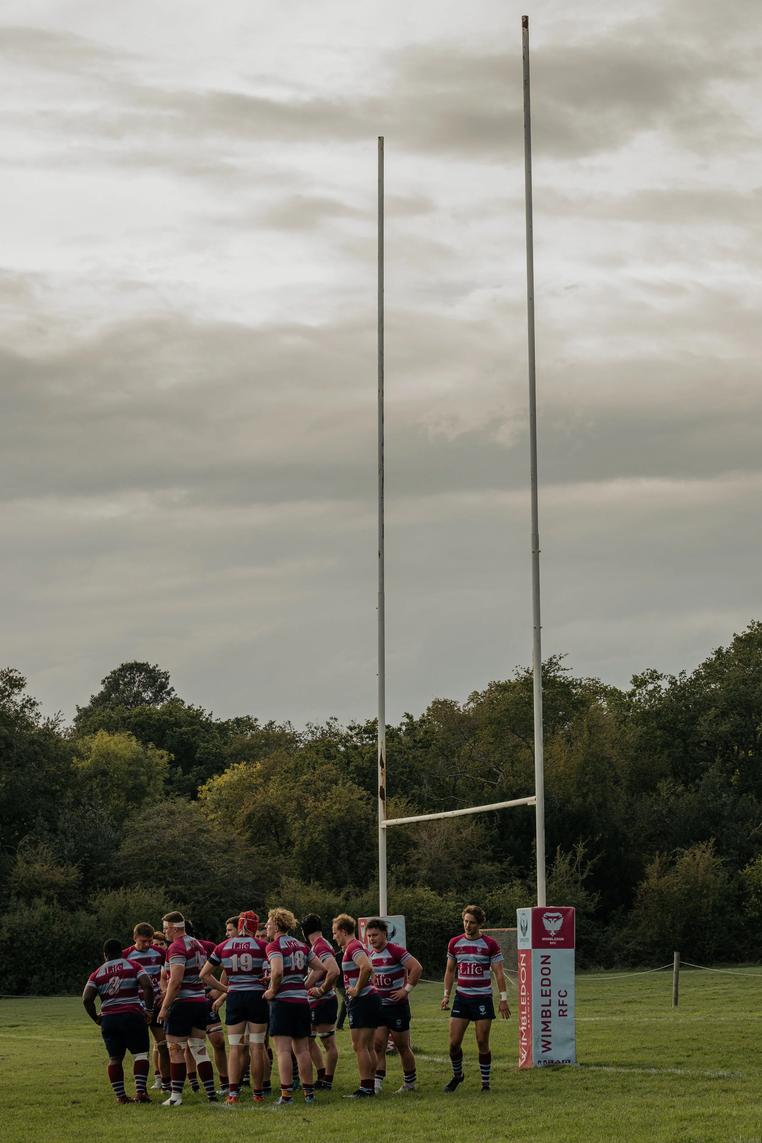 Rugby players in maroon and blue uniforms standing on a field near goal posts, with a sign for Wimbledon RFC, under an overcast sky, surrounded by trees.