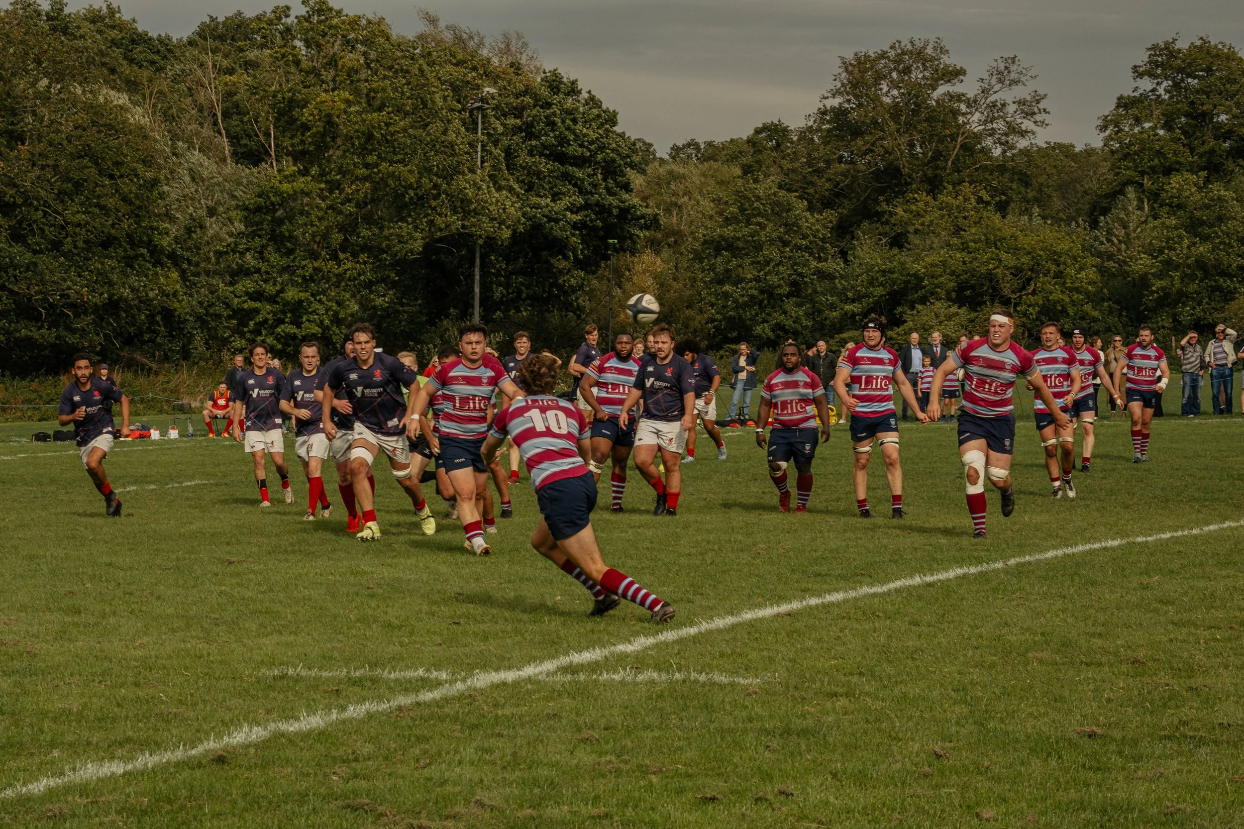 Rugby players in action during a game on a grassy field, with spectators watching in the background and trees surrounding the field.