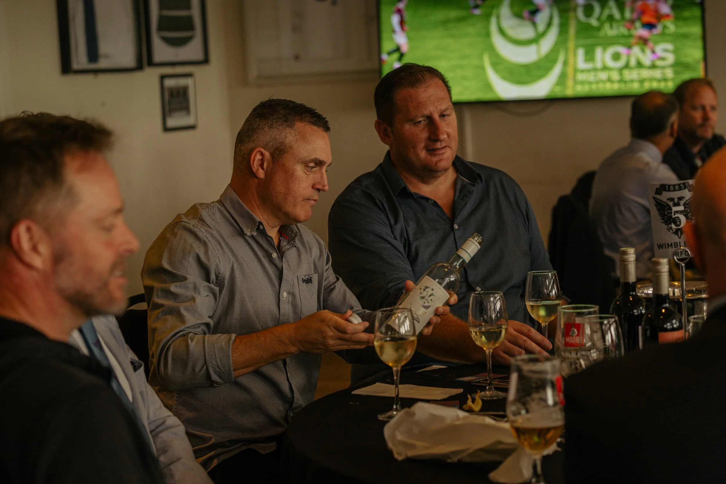 Men sitting at a table with wine glasses, one man holding a bottle of white wine, in a dimly lit restaurant or event space, with a sports game playing on a TV in the background.