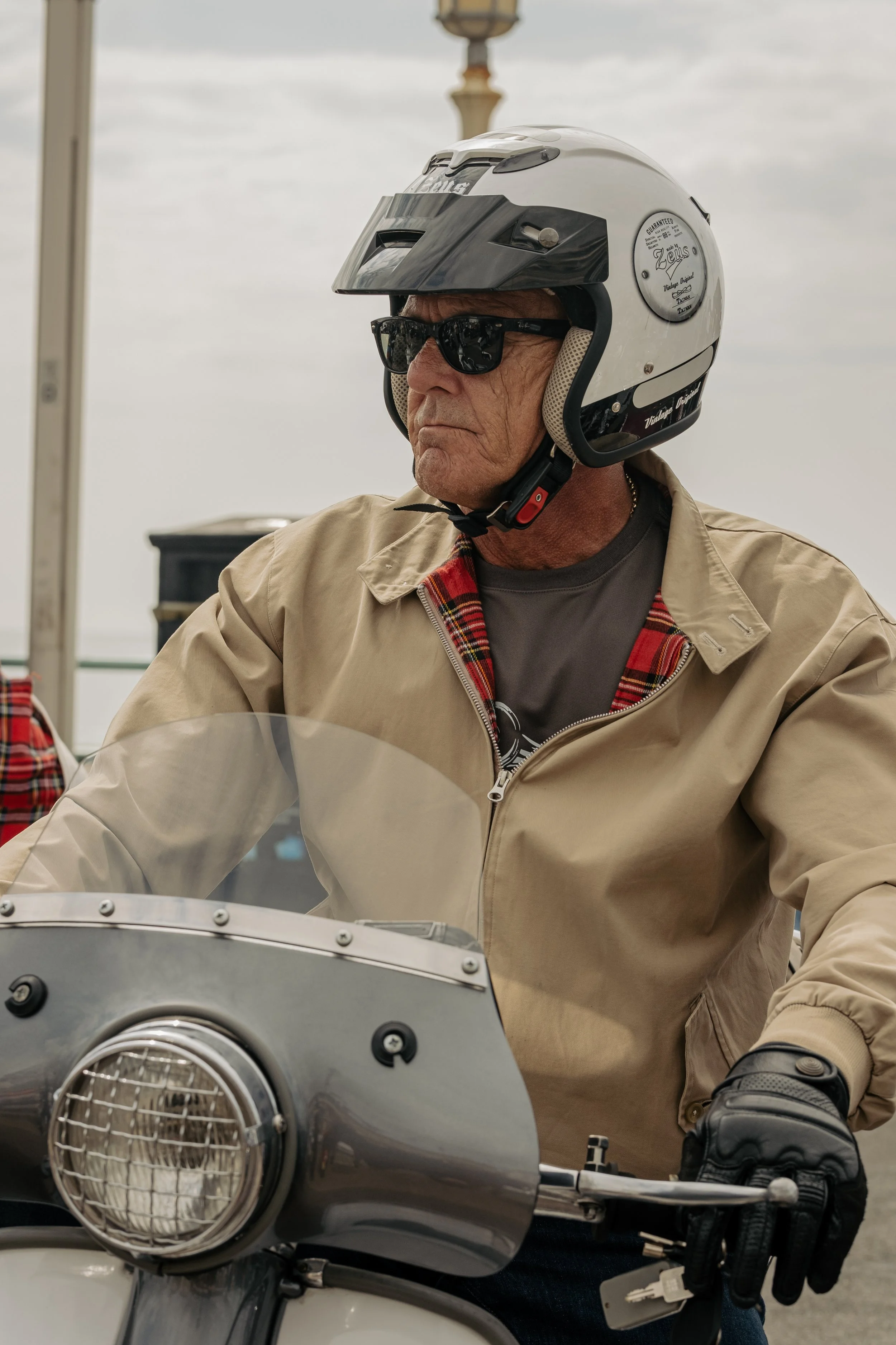An older man wearing a white helmet, sunglasses, a tan jacket, and black gloves sits on a motorcycle, riding on a cloudy day.