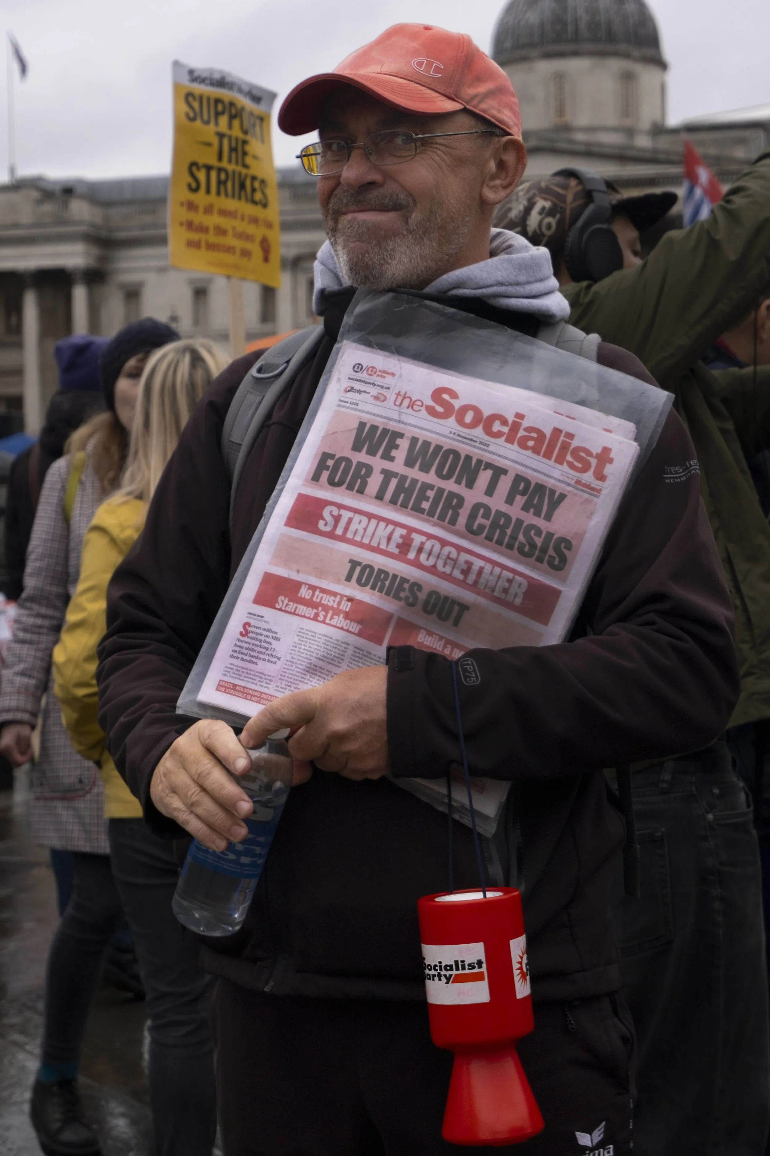 A man at a protest holding a newspaper with the headline 'We won't pay for their crisis' and a folder. He is wearing glasses, a red cap, and a black jacket. Several people and a building are in the background, along with a yellow sign supporting stri