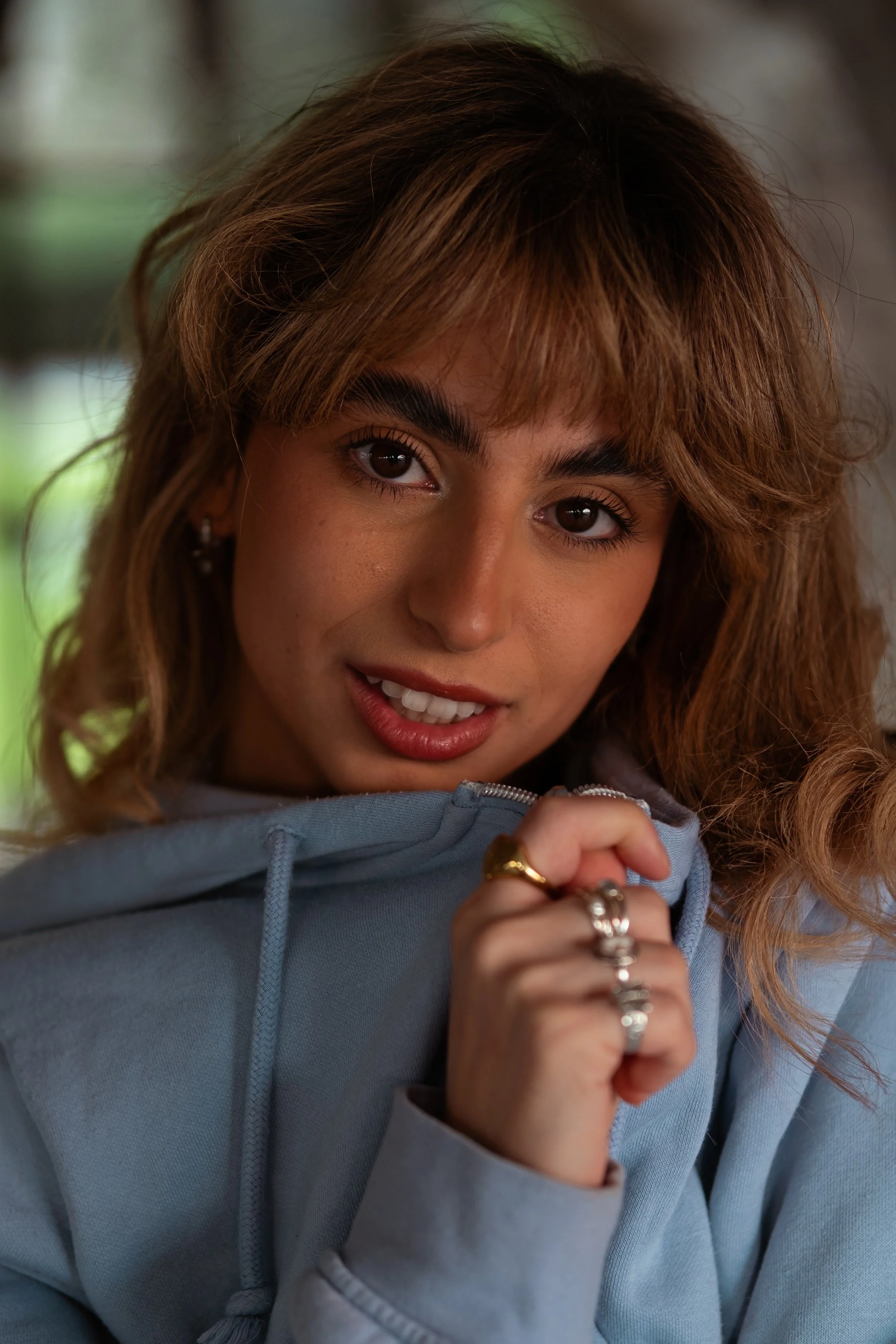 Close-up of a young woman with curly light brown hair, smiling slightly, wearing a light blue hoodie and rings on her fingers, looking into the camera.