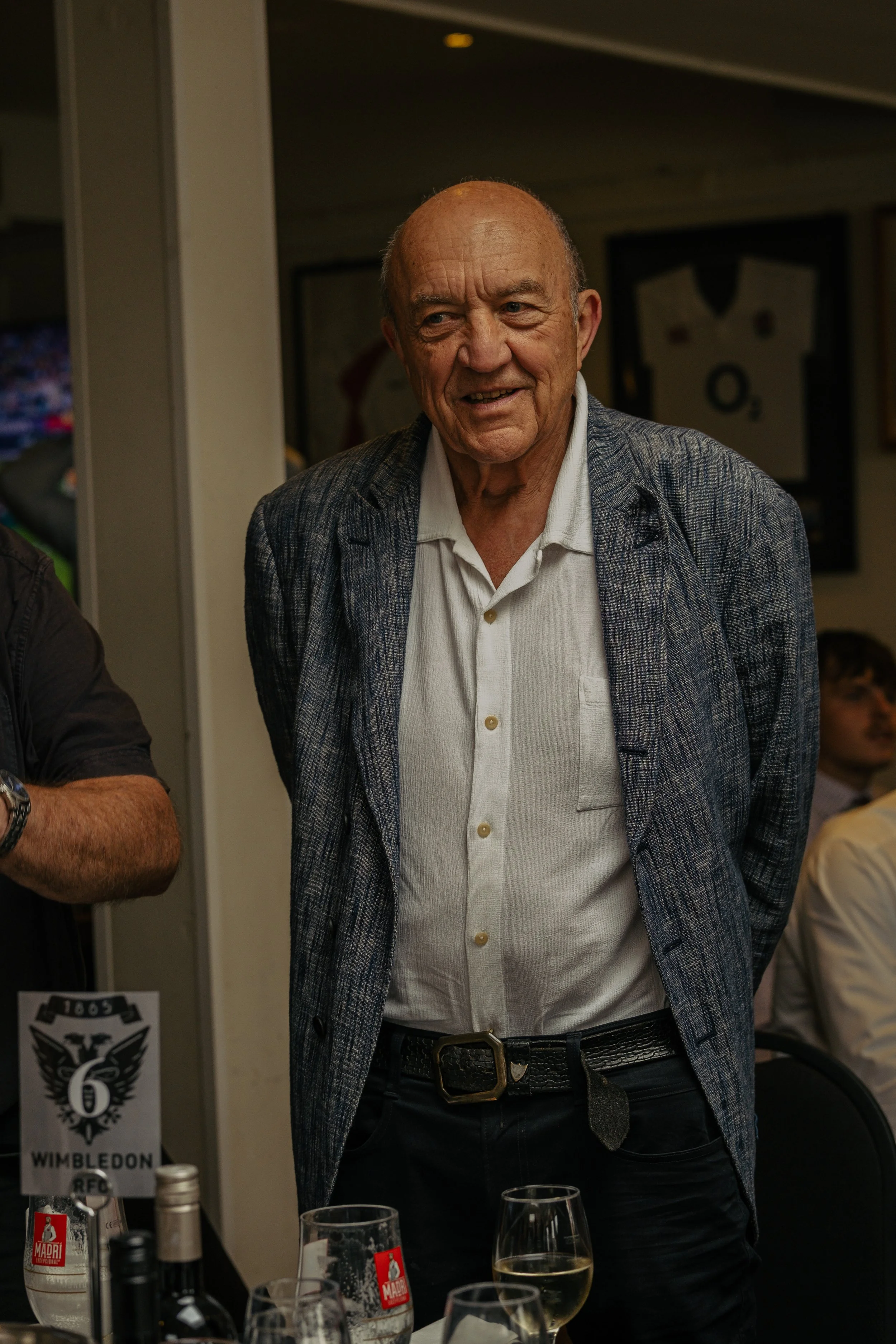 An elderly man with a bald head smiling during a social event, wearing a white shirt and a plaid blazer, standing behind a table with wine glasses and bottles.