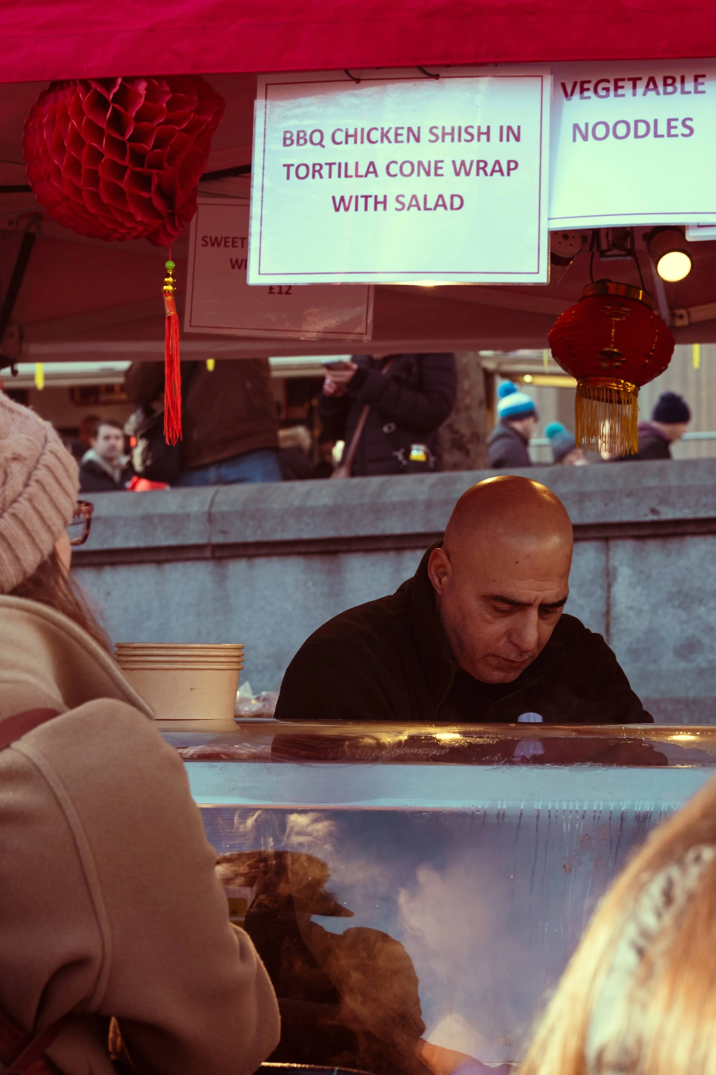 Bald man sitting at a food stall with a sign above advertising BBQ chicken shish in tortilla cone wrap with salad, surrounded by people at an outdoor market.
