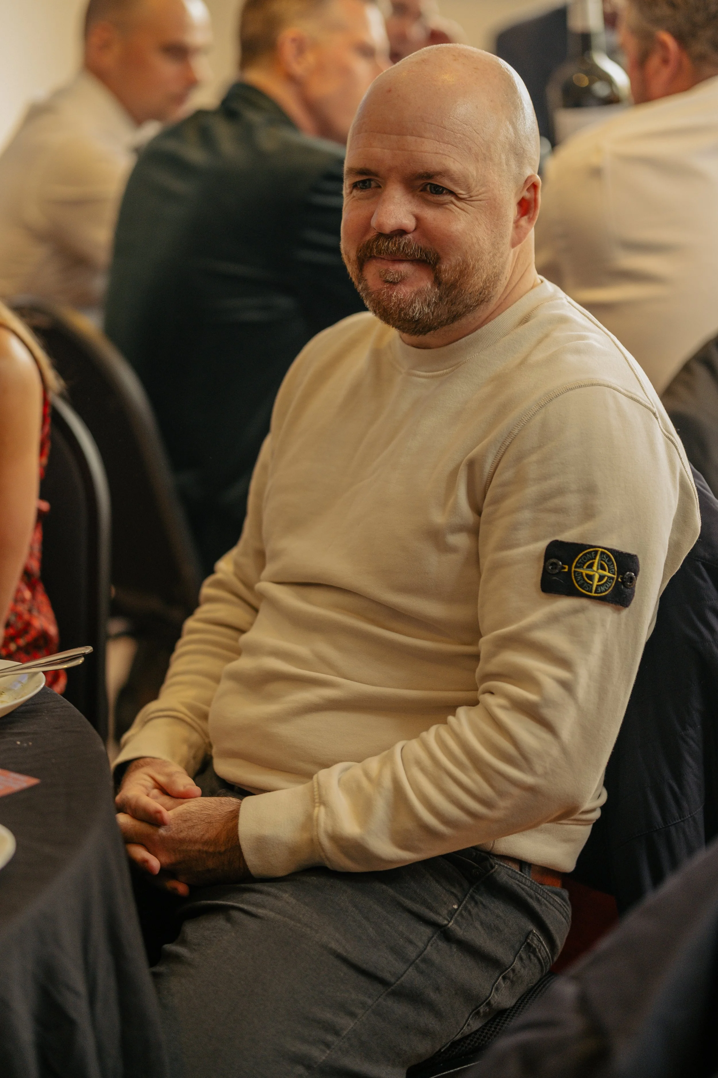 A man sitting at a table during a gathering or meeting, wearing a beige sweatshirt with a logo patch on the sleeve, and smiling while looking at the camera.