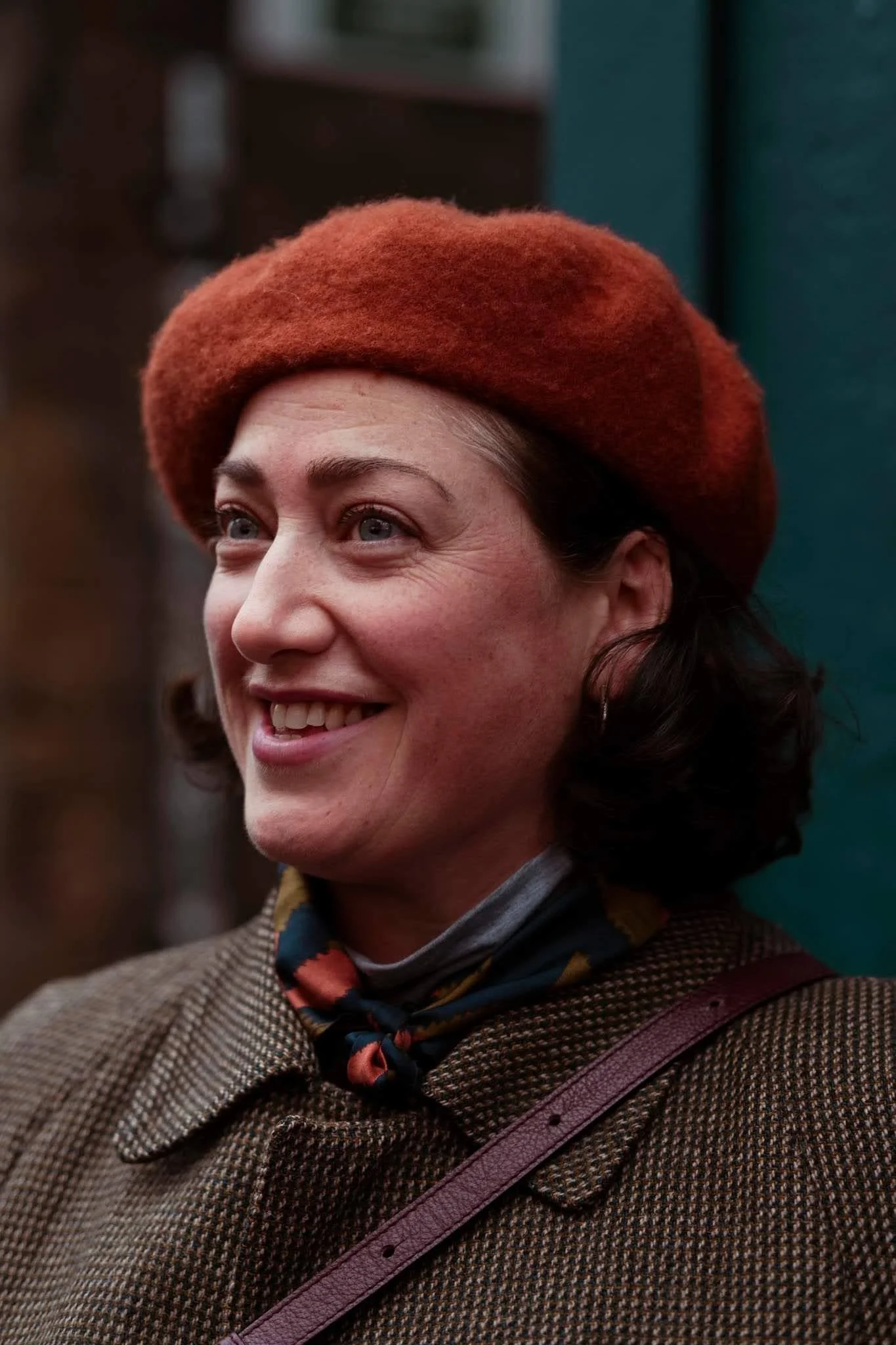 A woman wearing a brown coat, patterned scarf, and red beret smiling outdoors.
