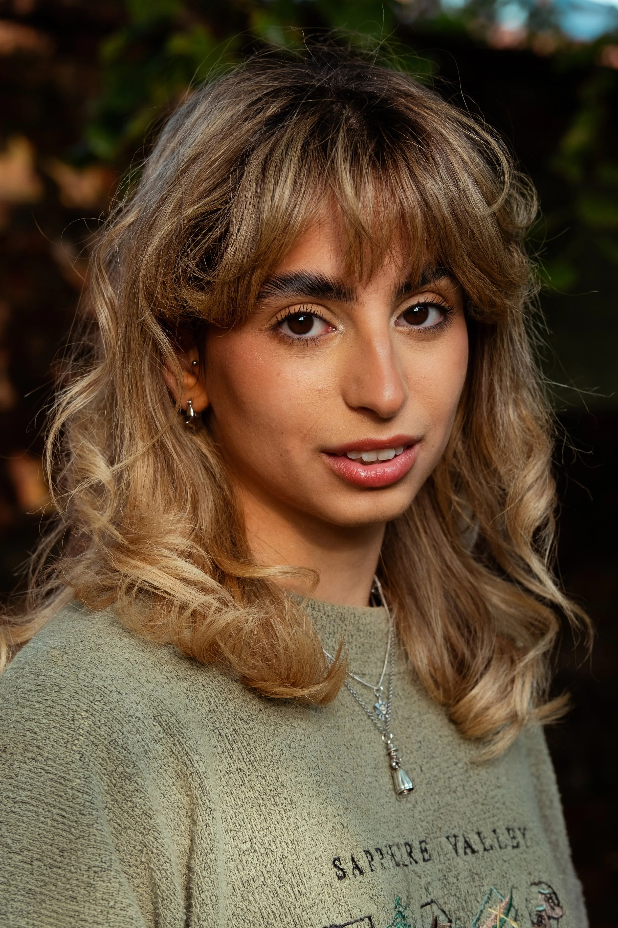 Close-up of a young woman with wavy light brown hair, wearing a green sweatshirt and layered necklaces, standing outdoors with blurred greenery in the background.