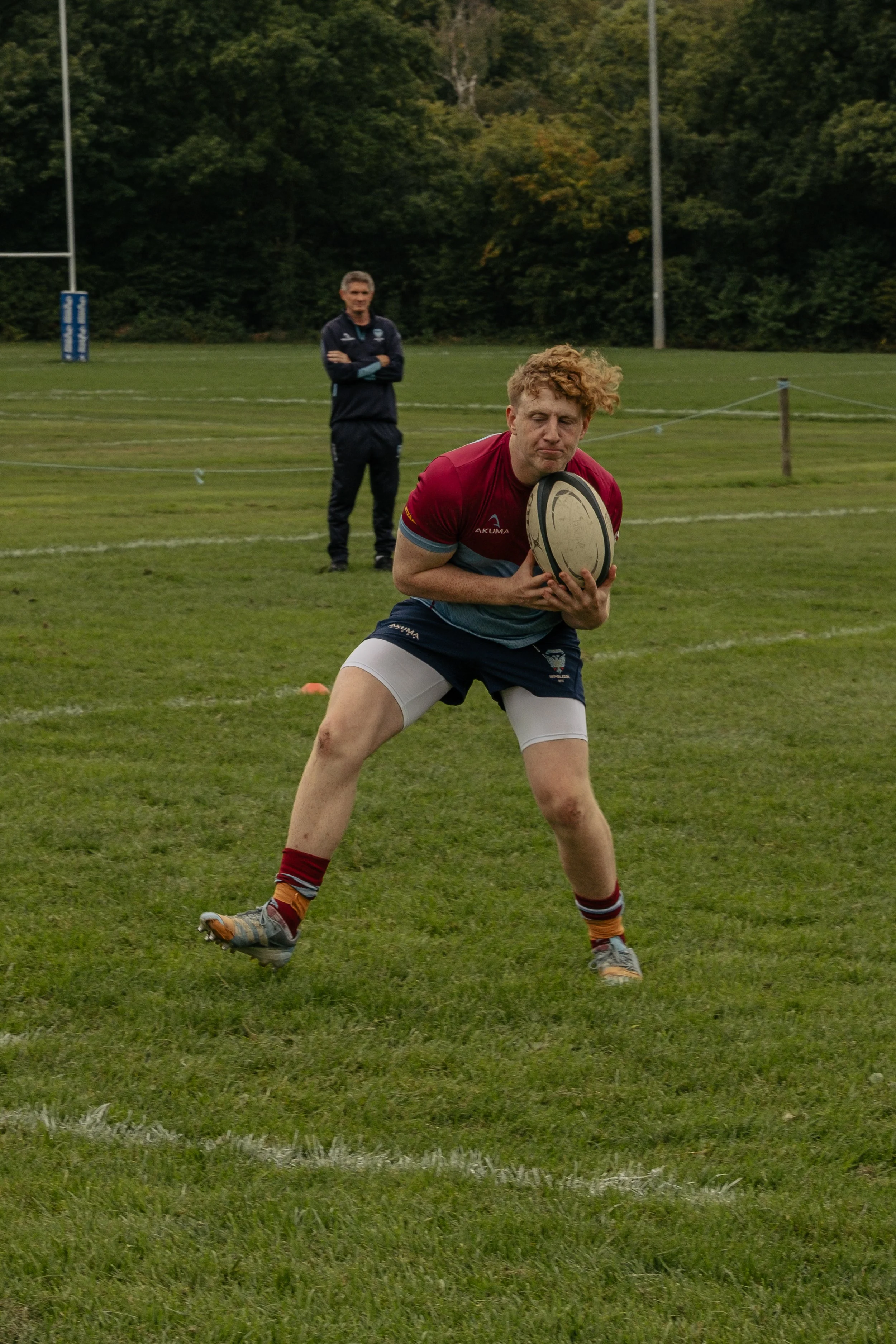 A rugby player holding a rugby ball on a grassy field, with a coach standing in the background, surrounded by trees and rugby goal posts.