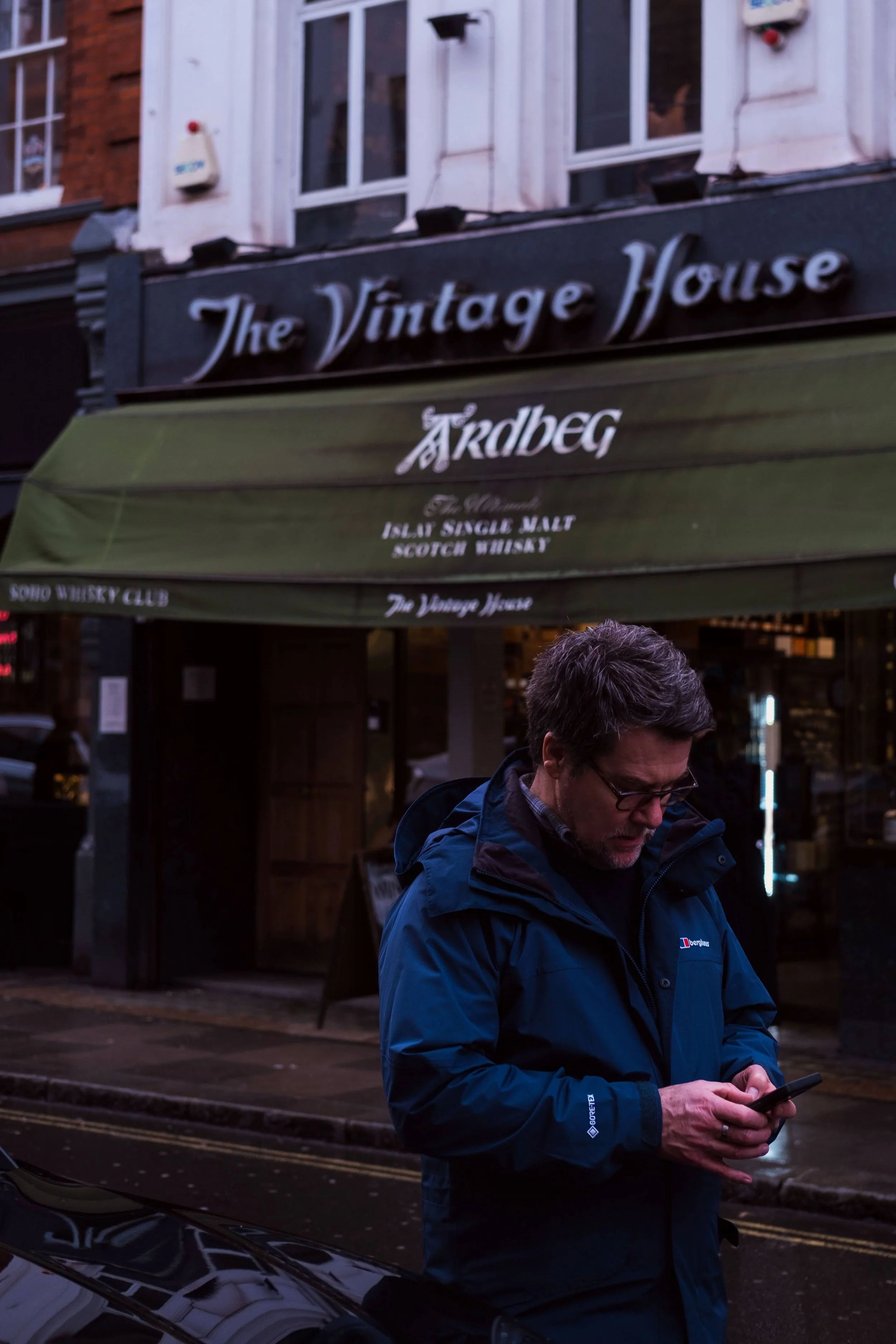 A man wearing a blue jacket and glasses looking at his phone on a rainy street in front of a bar called The Vintage House.