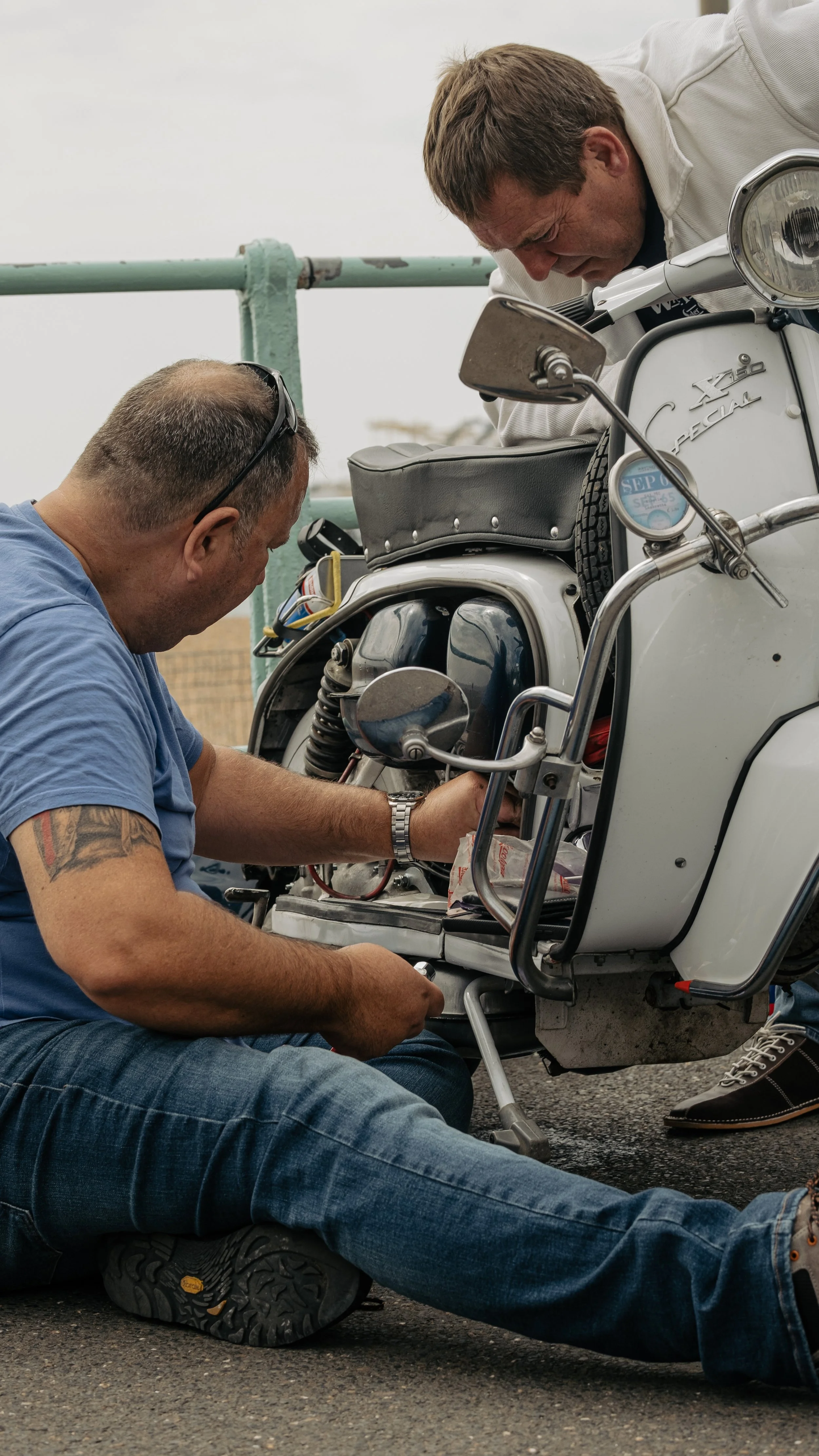Two men working on a white vintage scooter, with one sitting on the ground and the other leaning over the scooter's seat, making repairs.