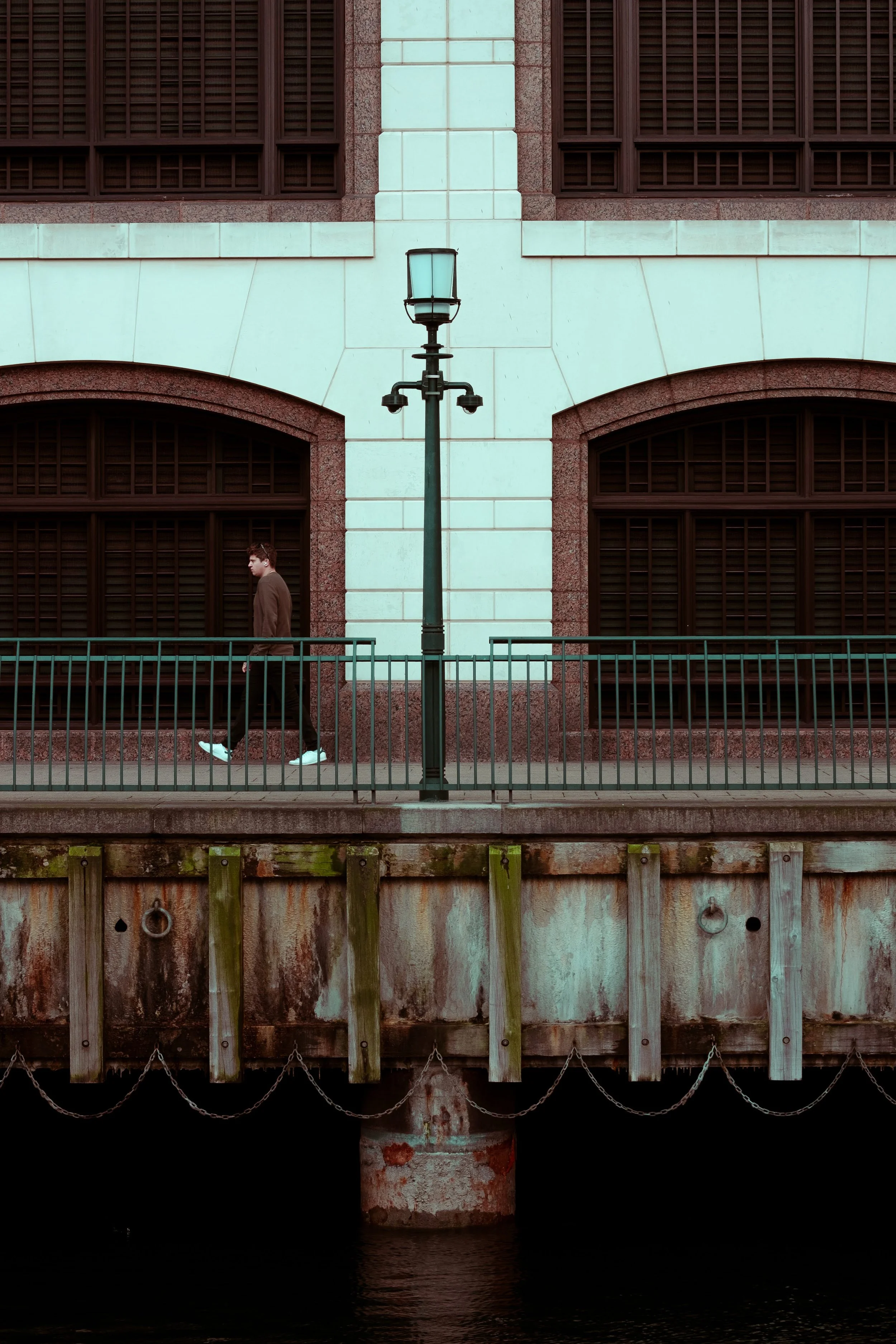 A man walking along a waterfront promenade with a lamppost and large windows in the background.
