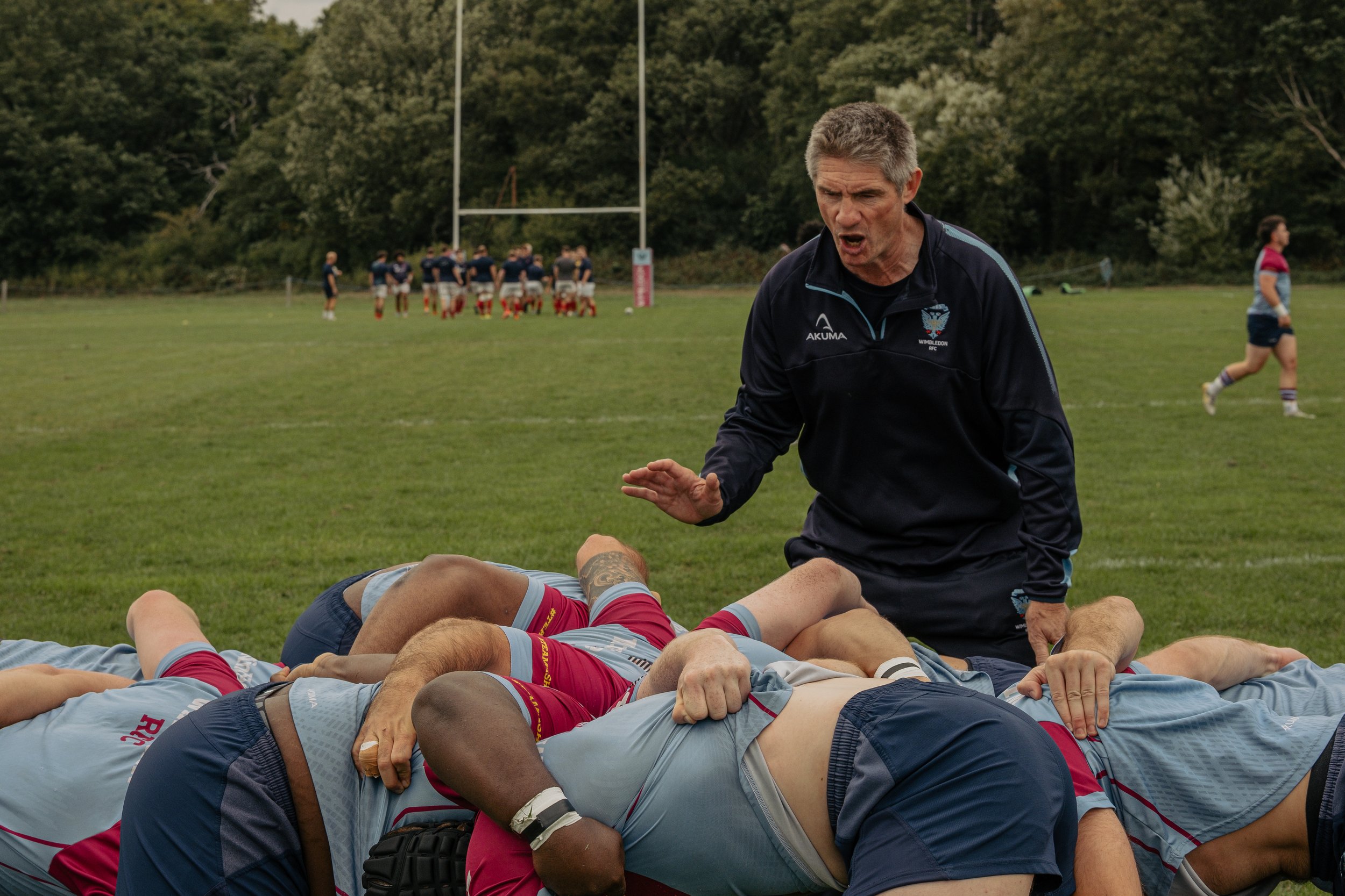 A rugby coach giving instructions to players lying on the ground during a game on a grassy field, with other players and a goalpost in the background.