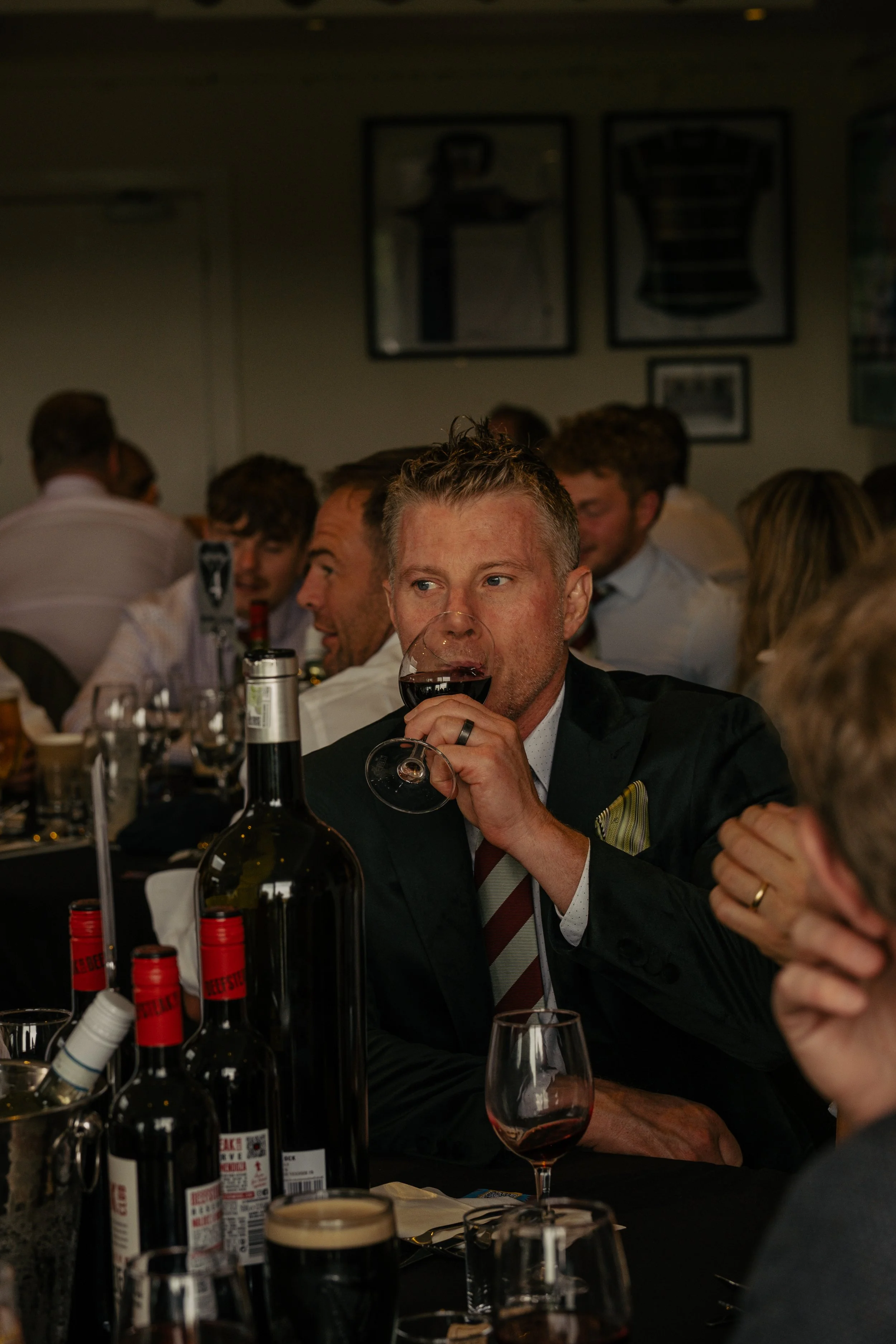 A man in a suit and striped tie drinking red wine at a dinner event, surrounded by other people at a large table with bottles, glasses, and tableware.