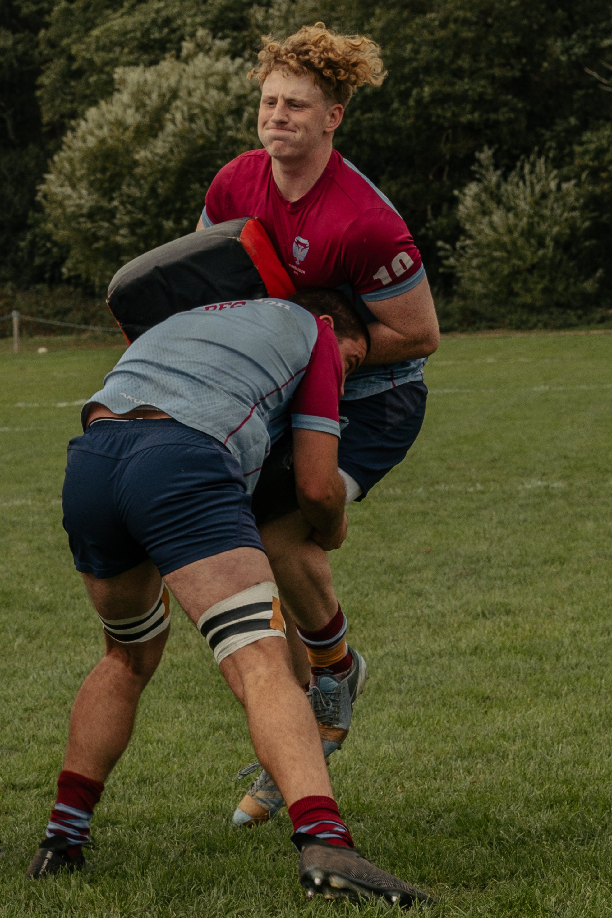 Two rugby players are engaged in a tackle during a game on a grassy field, with trees in the background.