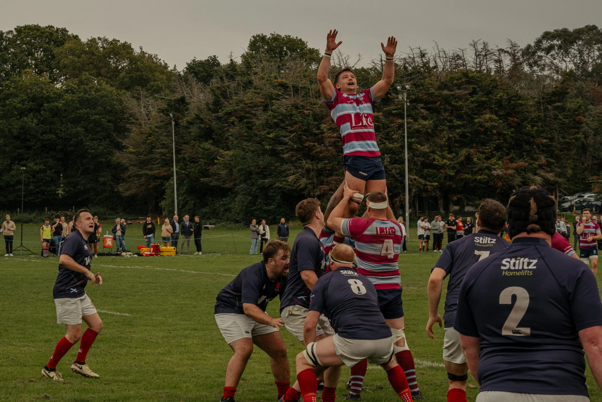 Rugby players performing a line-out during a match, with one player lifted into the air and reaching for the ball, on a grassy field with spectators and trees in the background.