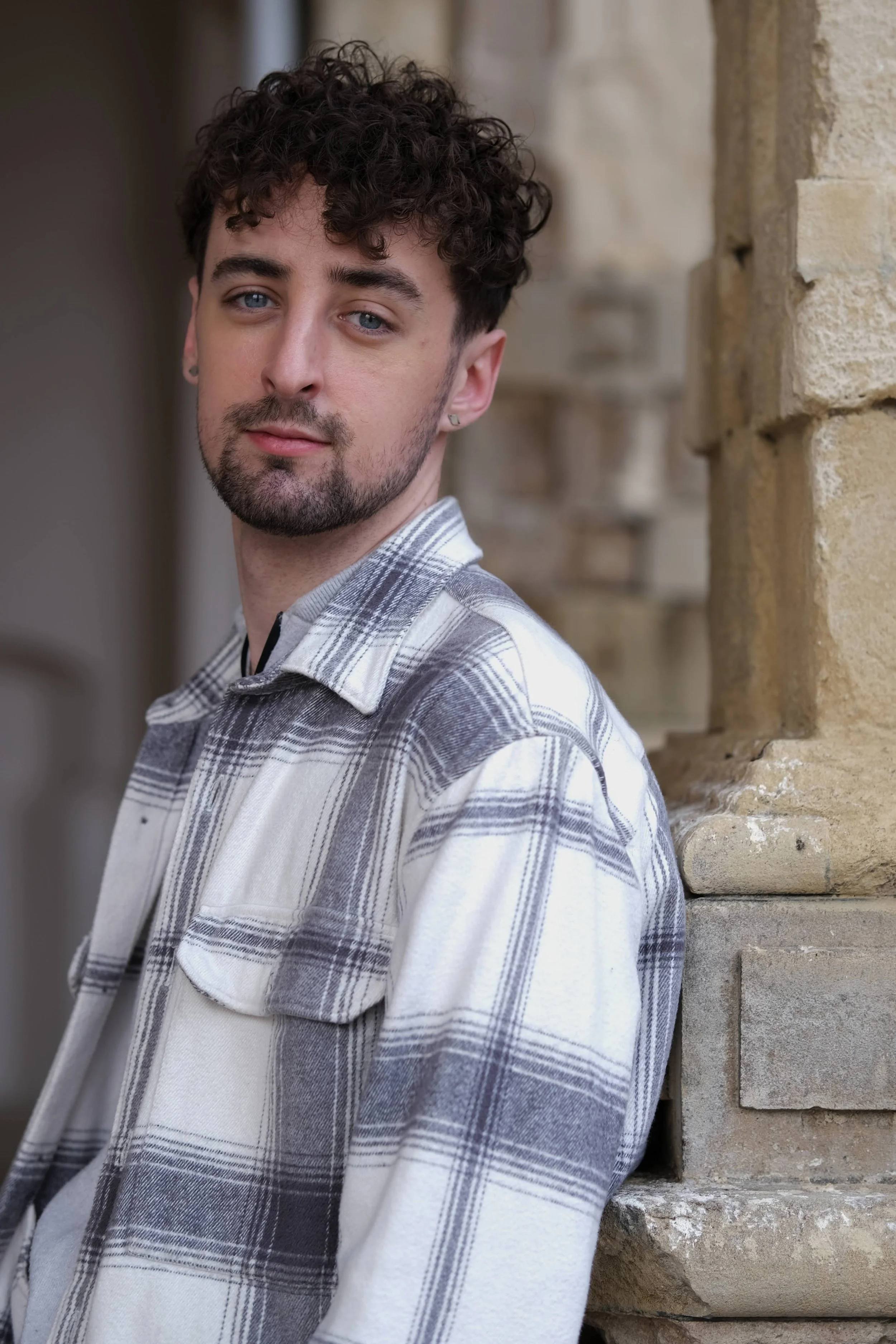 A young man with curly dark hair, blue eyes, and a beard leaning against a tan brick wall, wearing a white and gray plaid shirt, looking at the camera.
