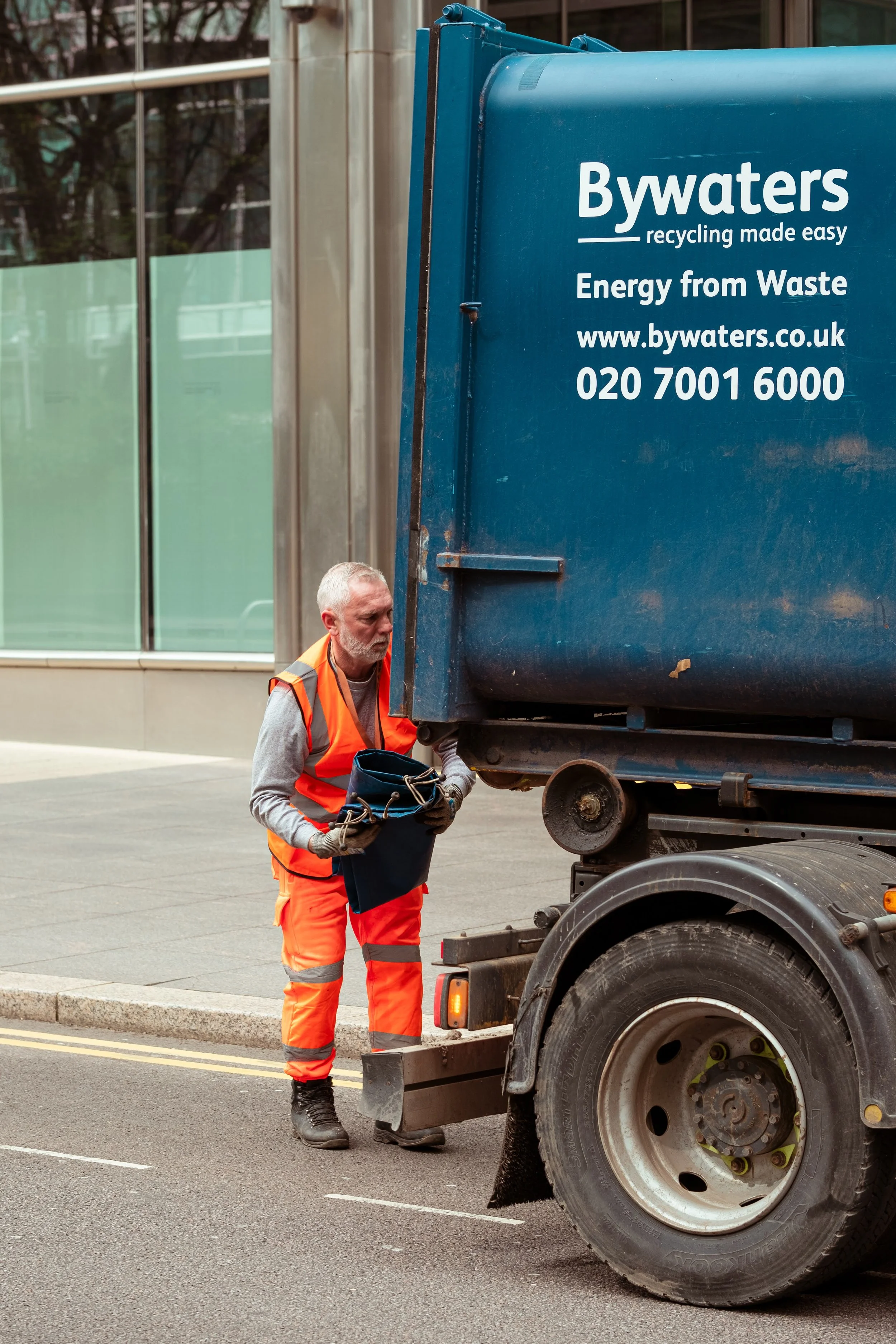 A sanitation worker in an orange reflective vest and pants inspecting a blue waste collection truck on a city street.
