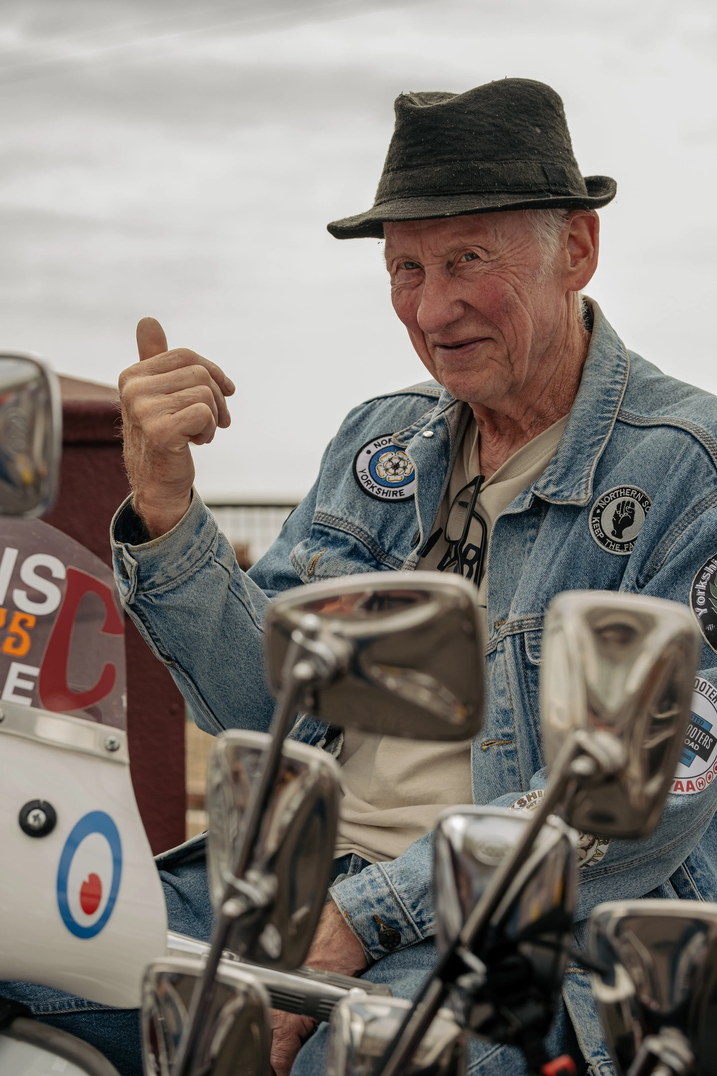 An elderly man with a cowboy hat and denim jacket sitting on a motorcycle, giving a thumbs-up and smiling.