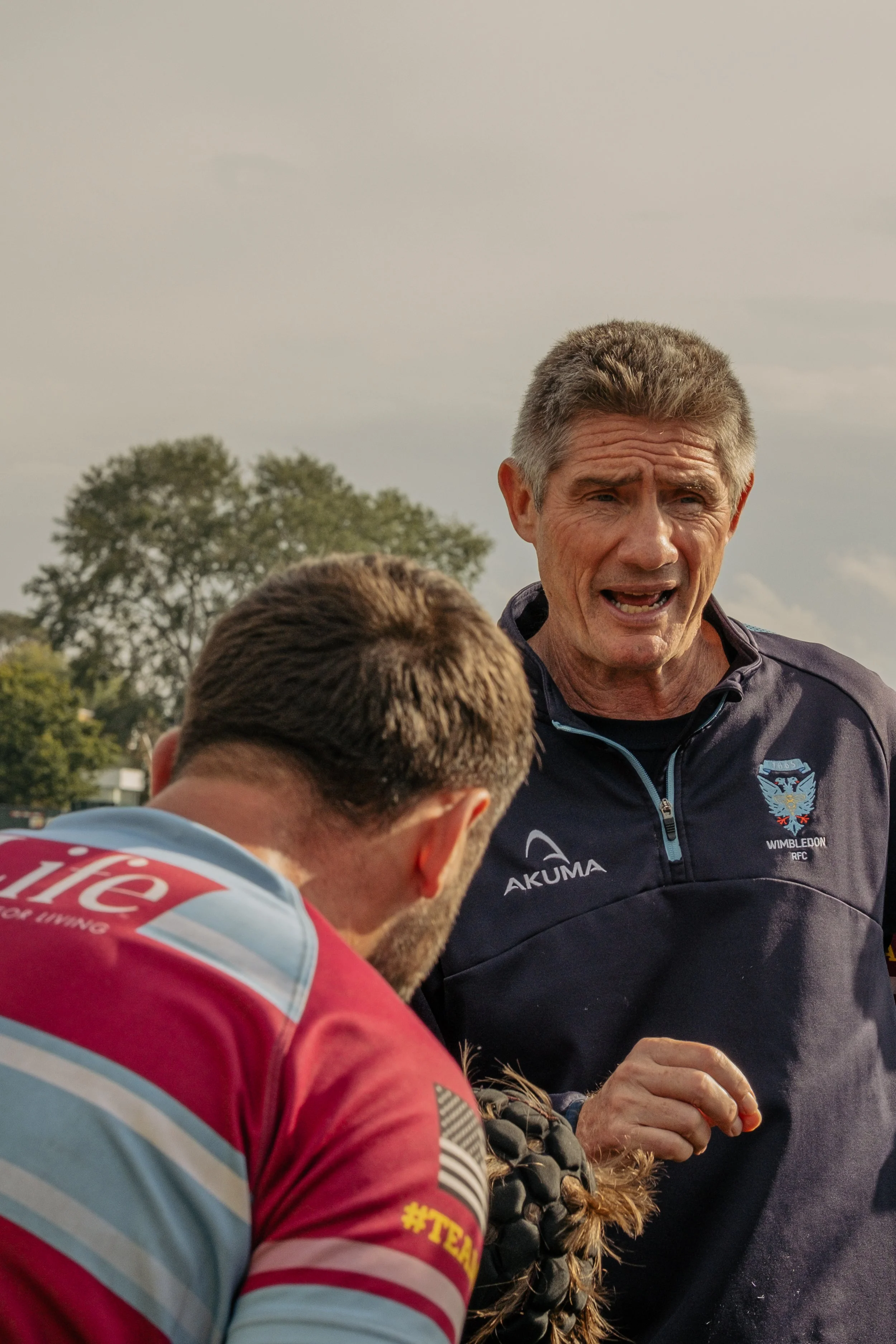 A man in a dark sports jacket with a Wimbledon logo and Akuma brand name is talking to another person, whose back is turned, during an outdoor sports event. A tree and cloudy sky are visible in the background.
