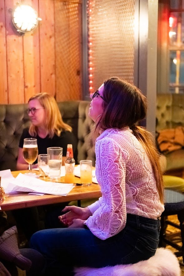 Woman in white lace top and glasses sitting at a table in a dimly lit restaurant or bar with drinks, with another woman in black sitting in the background.