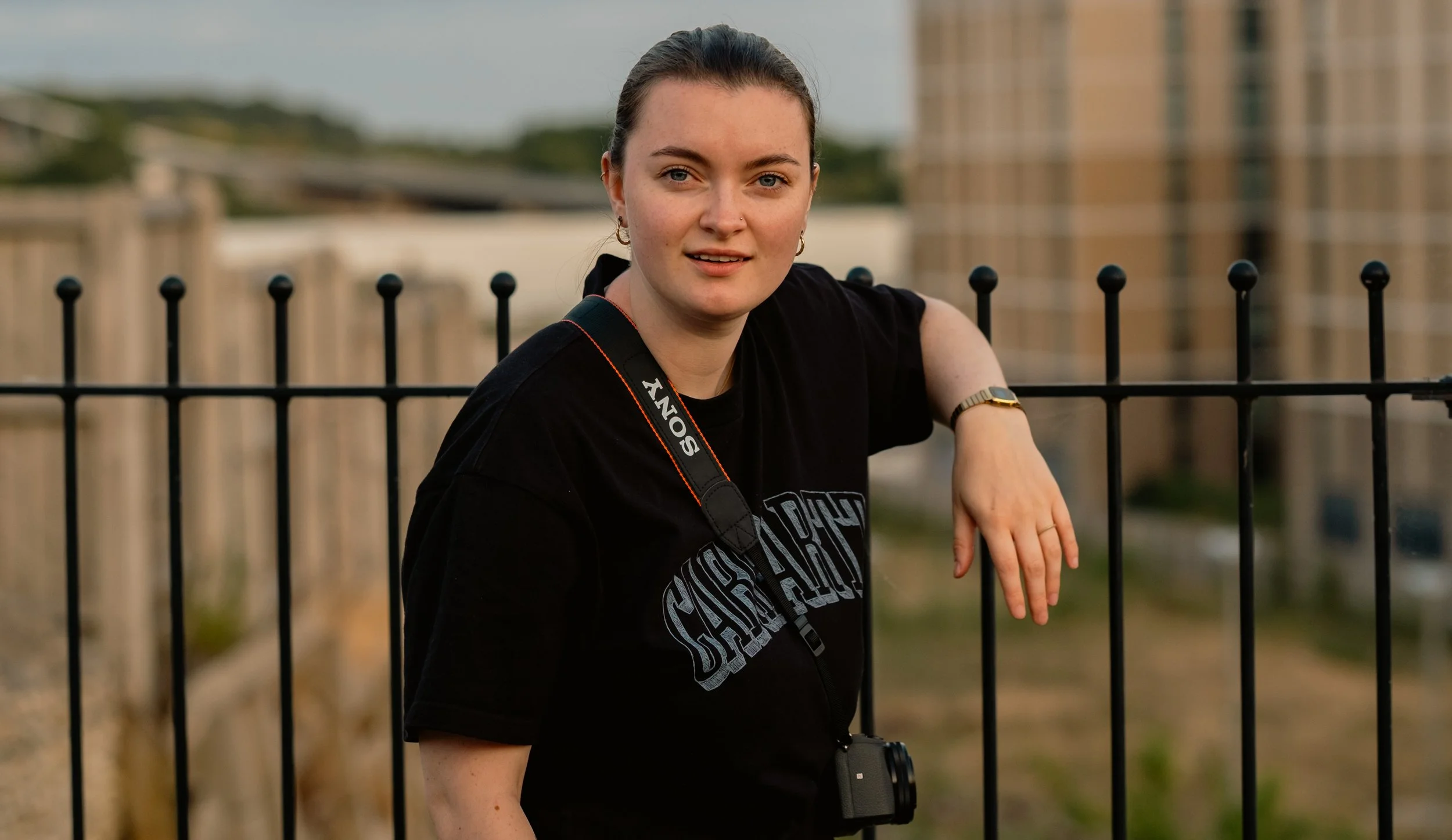 A young woman with brown hair, wearing a black 'CHISAR' T-shirt, a camera strap with 'SONY' written on it, and a watch, leaning on a black metal fence outdoors with a blurred cityscape in the background.