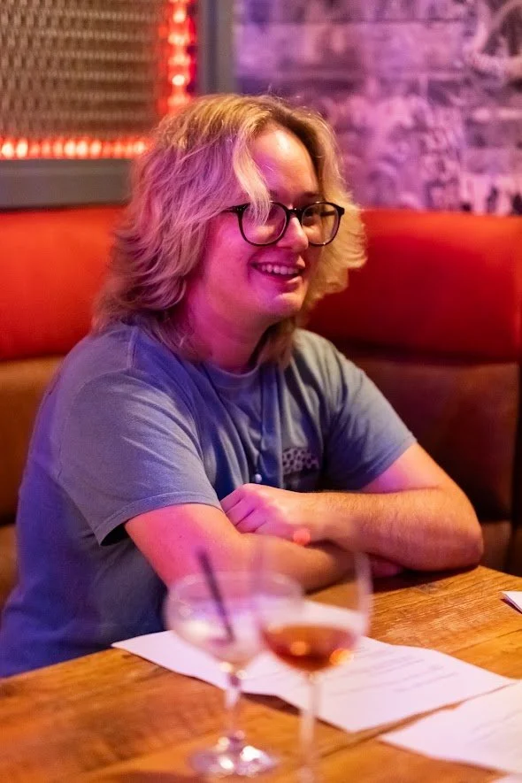 A person with blond, wavy hair and glasses smiling while sitting at a wooden table in a dimly lit bar or restaurant, with drinks and menus in front of them.