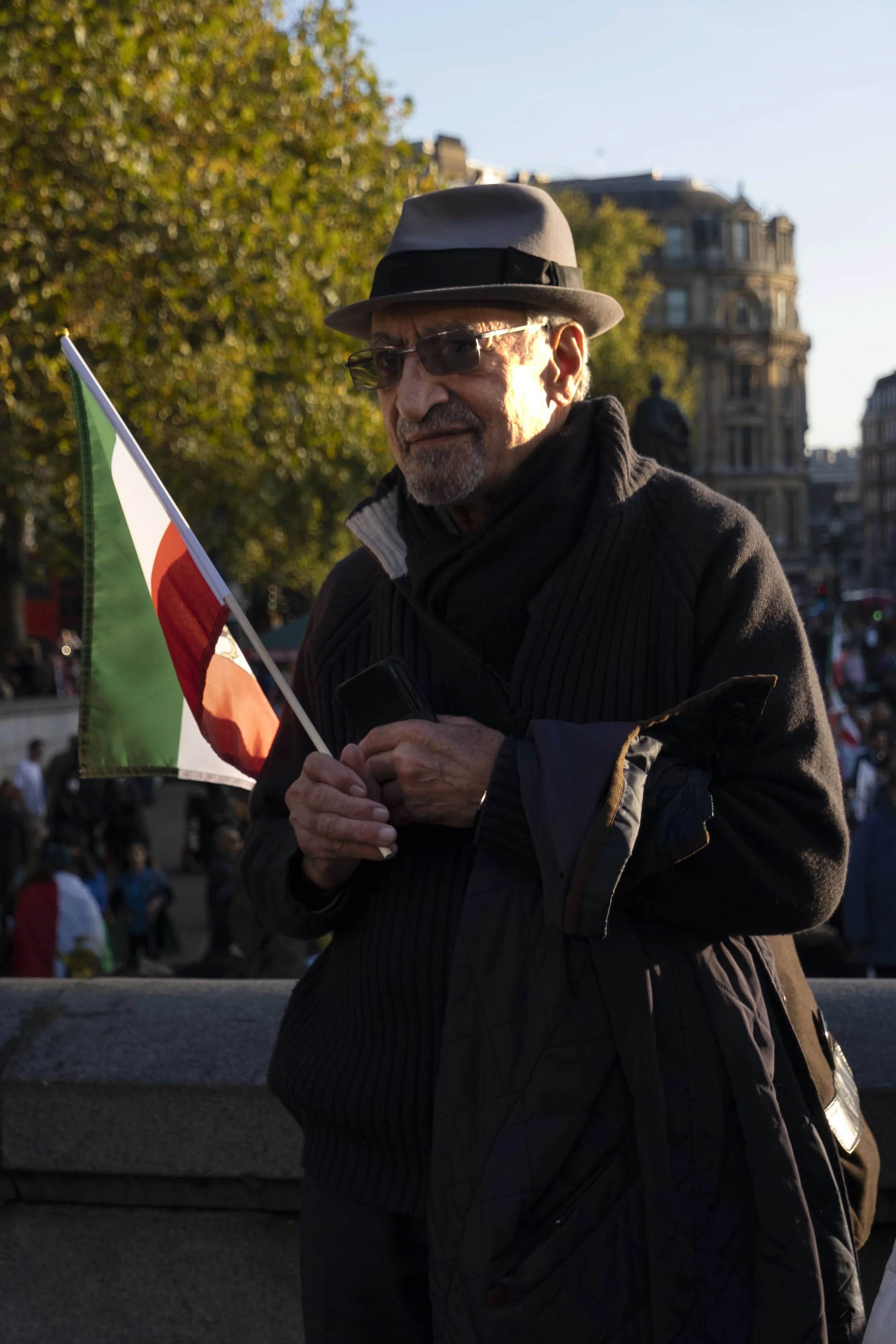 Older man wearing glasses, a hat, and a coat, holding a small Italian flag, standing outdoors in a city square with trees and historic buildings in the background.