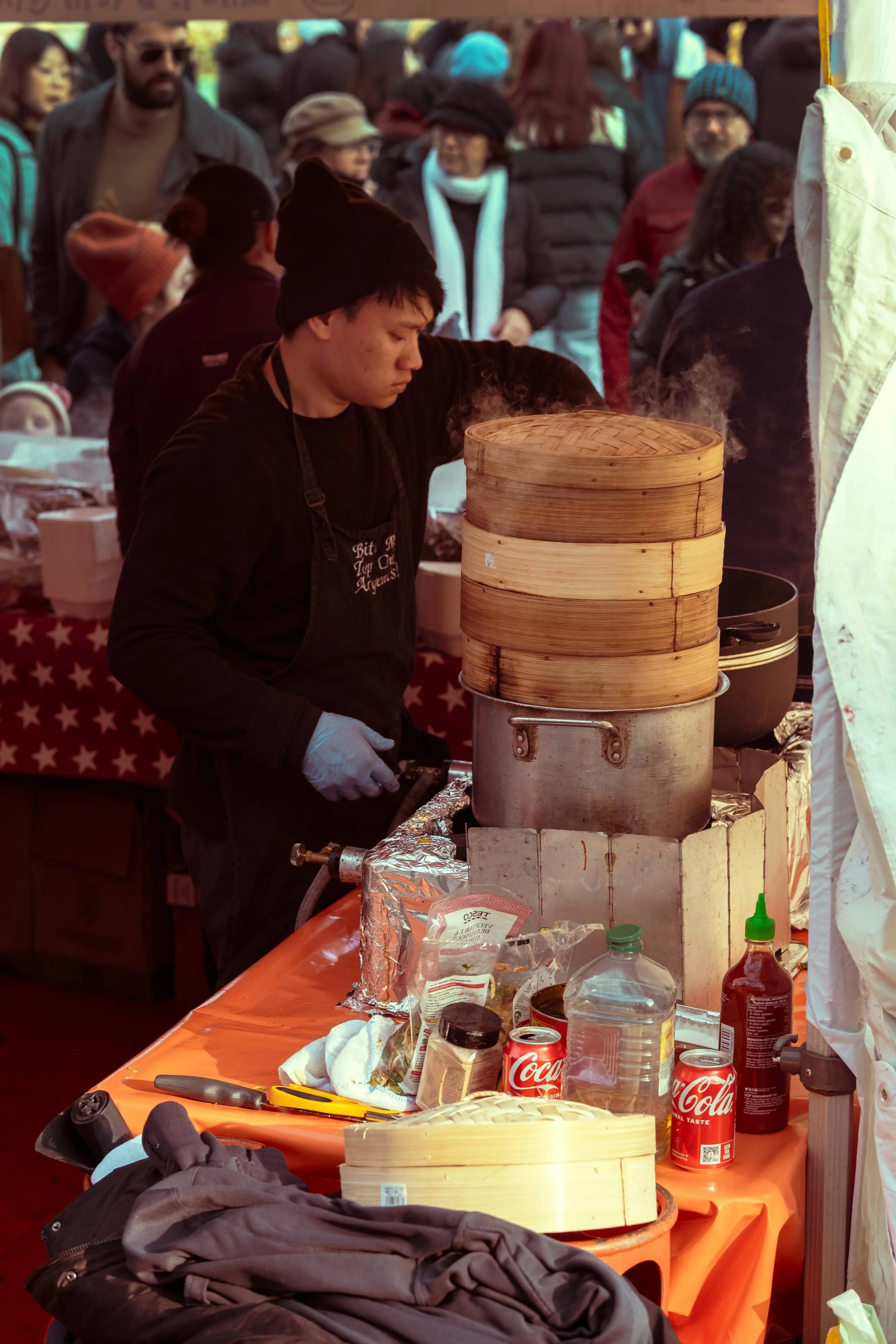 A young man making steamed dumplings at an outdoor food stall with a crowd in the background.