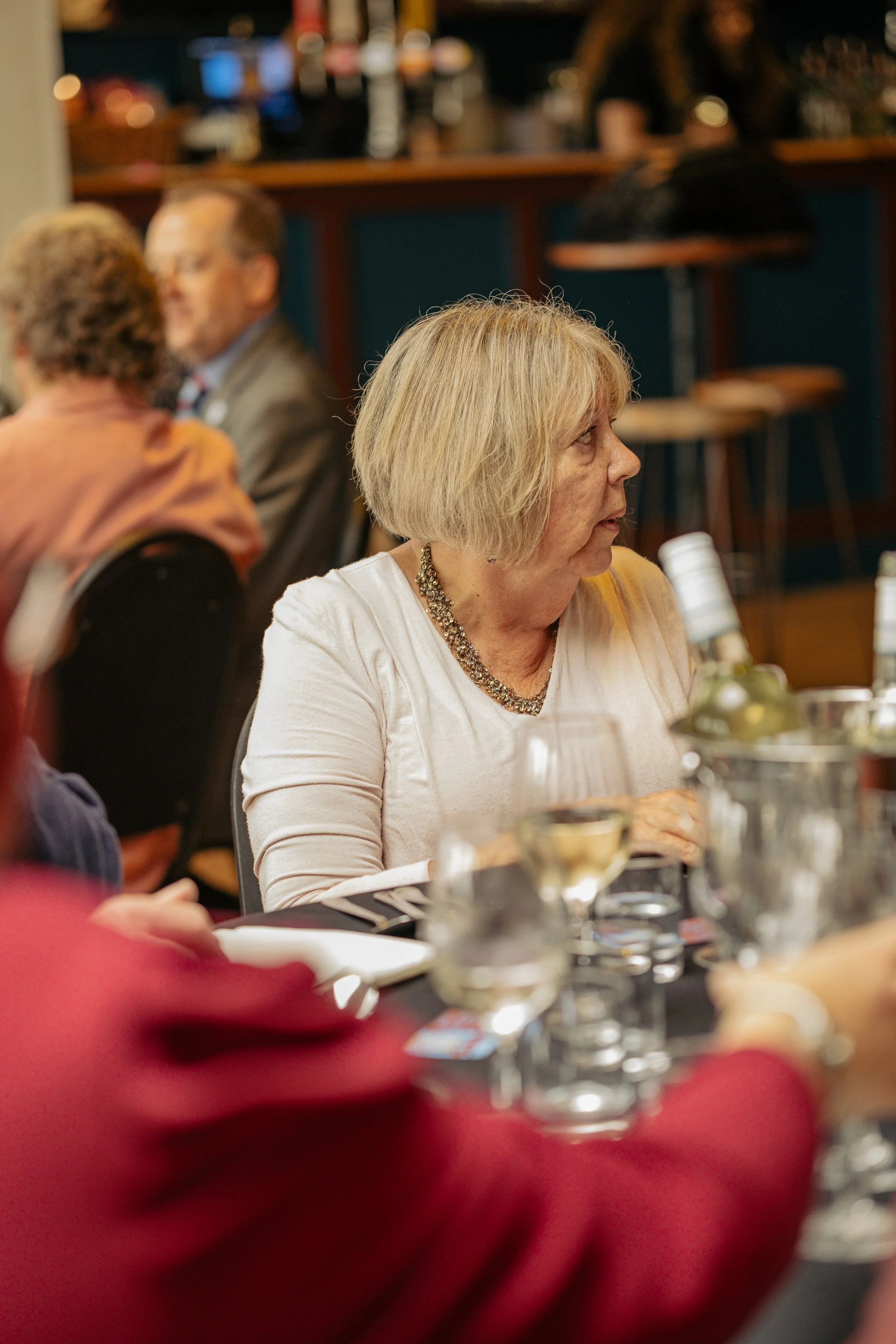 A woman with shoulder-length blonde hair and a necklace, sitting at a dining table with wine glasses and tables with other people in the background.