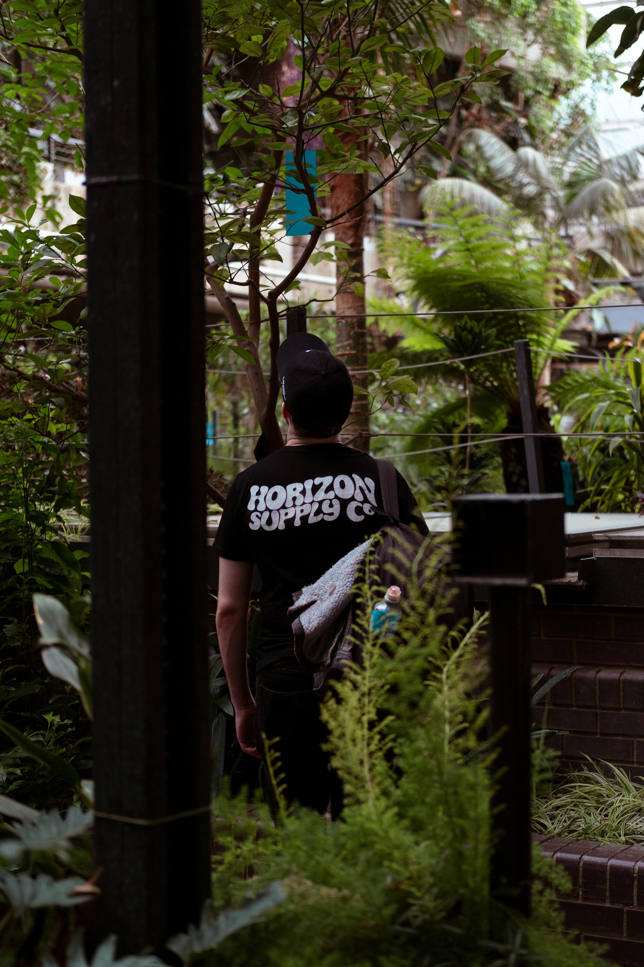 A person wearing a black t-shirt with 'Horizon Supply Co.' on the back, carrying a gray sweater and a water bottle, is walking through a lush, green garden or greenhouse filled with various plants and trees.