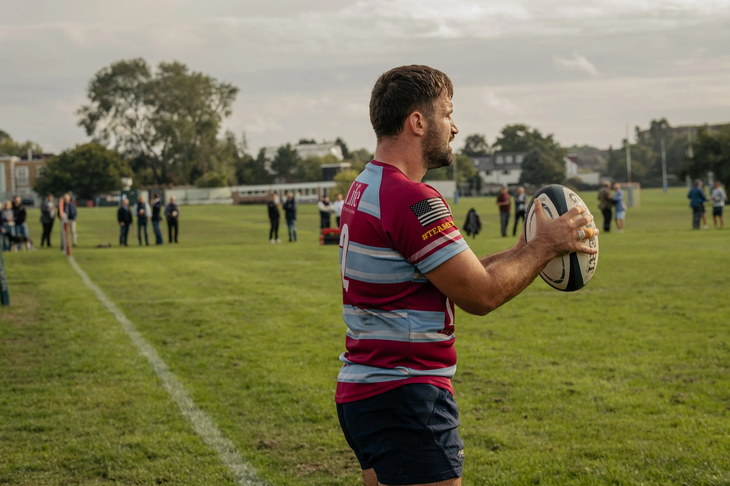 A man holding a rugby ball during a game or practice on a grassy field, with spectators or players in the background.