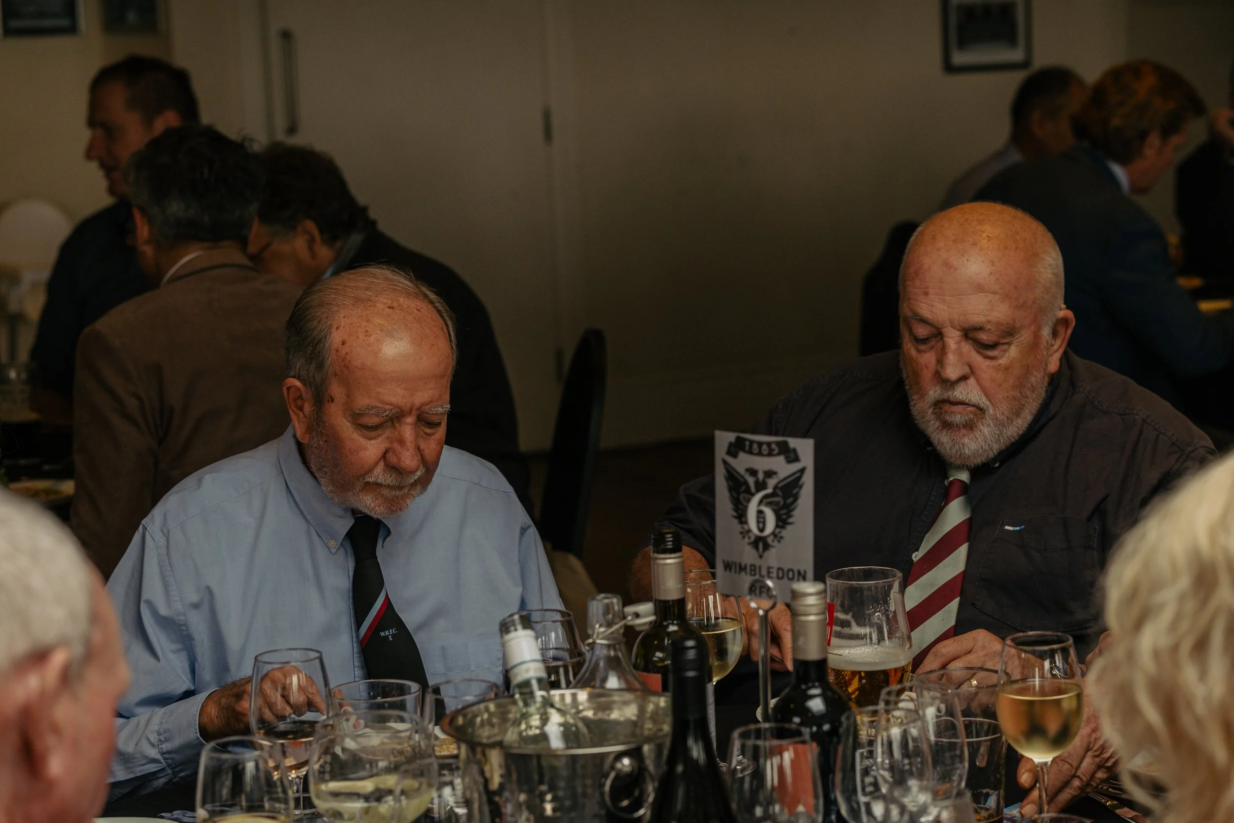 People seated at a dinner table, two elderly men in the foreground with drinks and bottles, a sign with the number 6 and the word Wembley, others blurred in the background.