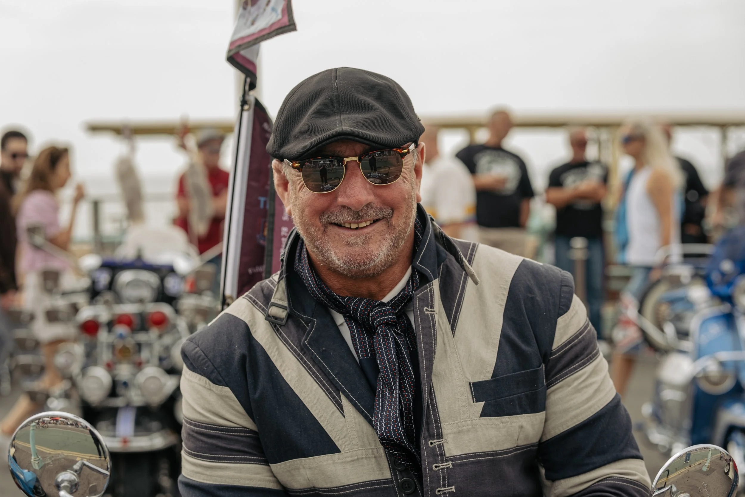 Smiling man wearing sunglasses, a black cap, and a vintage-style motorcycle jacket at a motorcycle gathering.