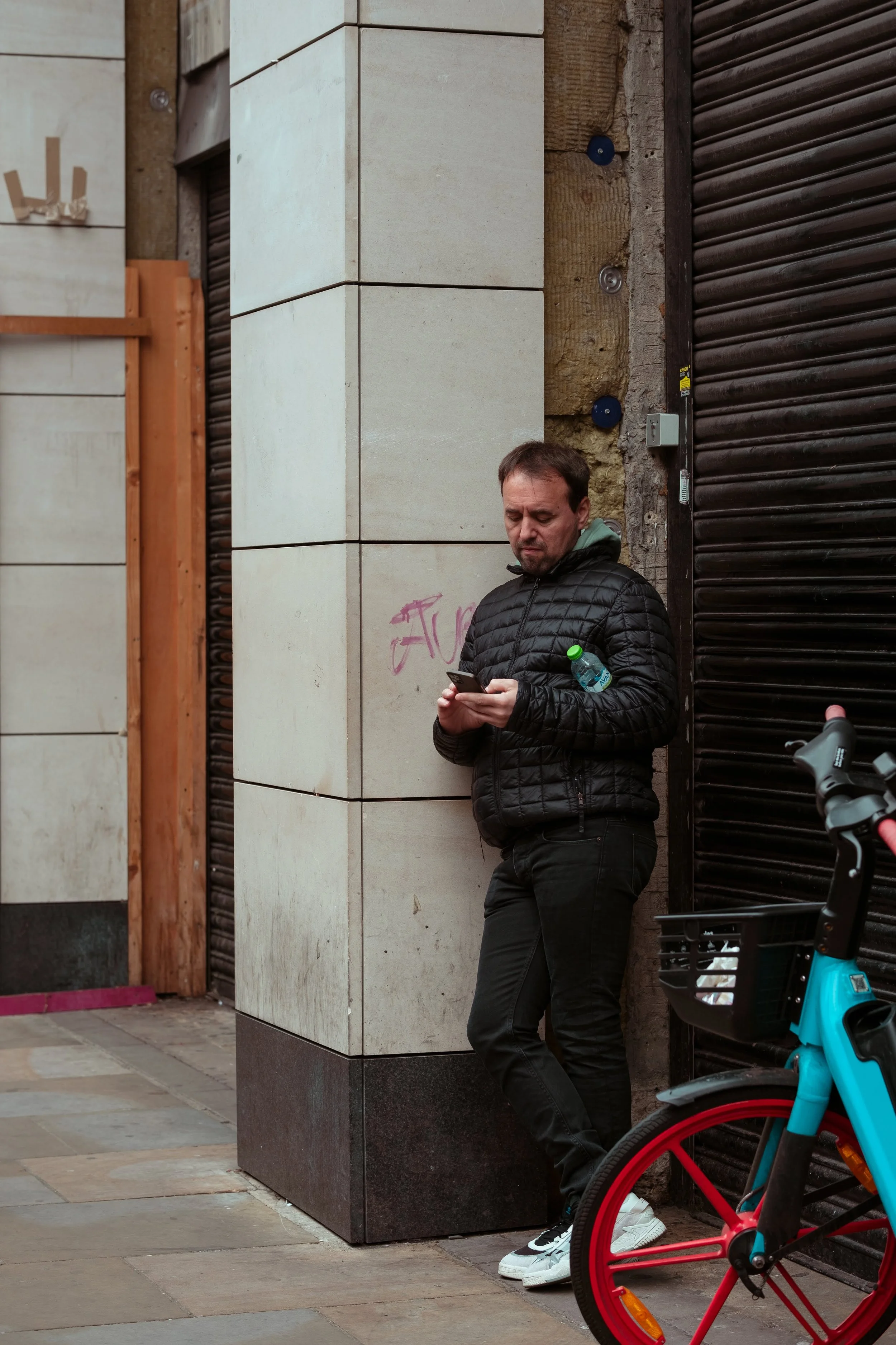 A man in a black jacket and black pants leaning against a wall in an urban area, looking at his phone, with a water bottle in his left pocket, and a blue bike with a black basket in front of him.