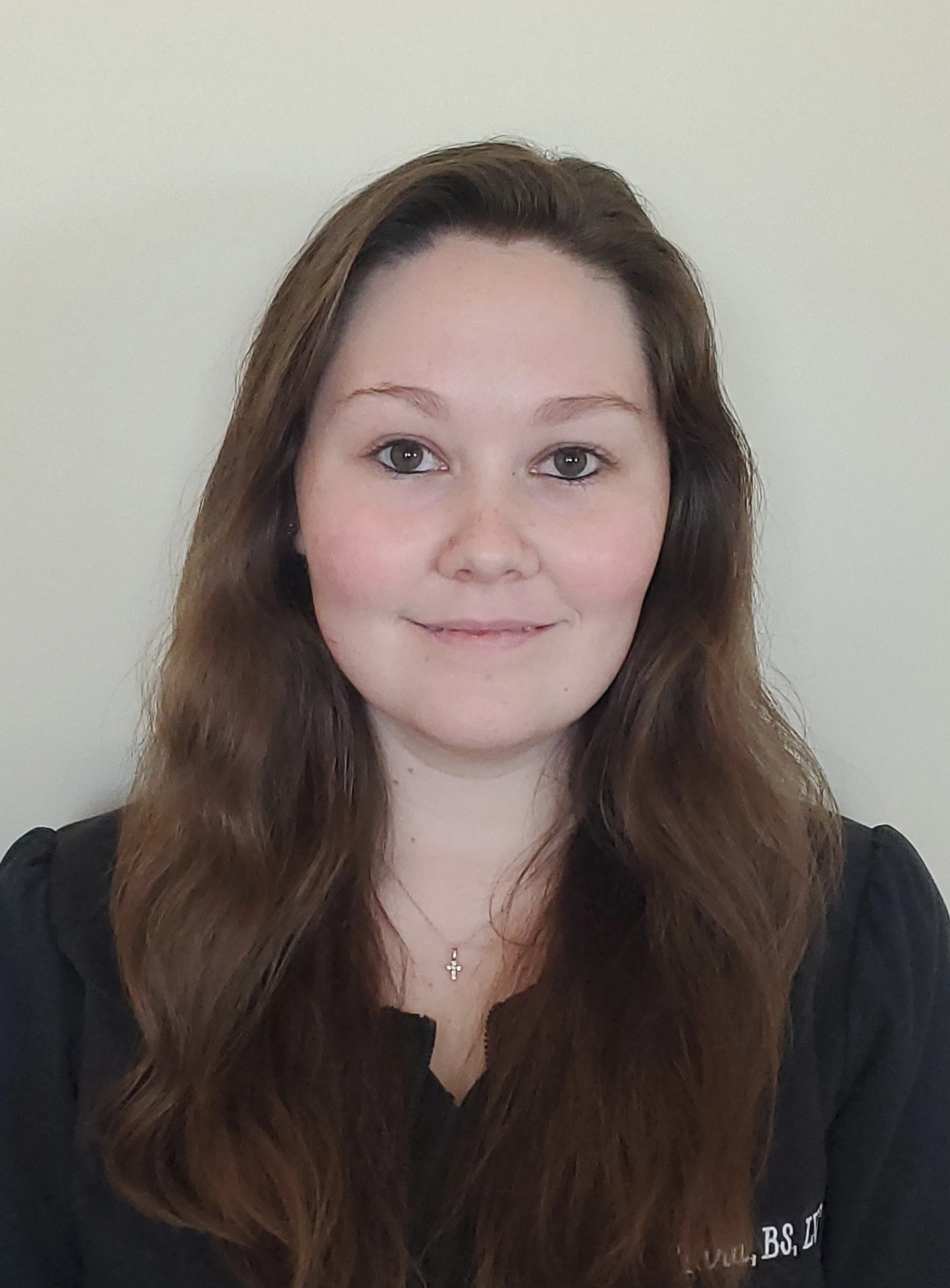 A young woman with long, wavy brown hair, fair skin, and gray eyes, smiling slightly, wearing a black top and a delicate cross necklace, against a plain light-colored background.