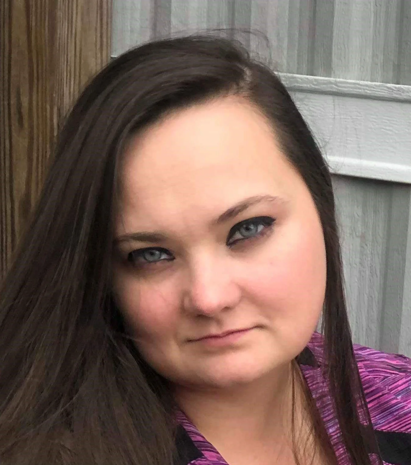 Close-up of a woman with long dark hair and blue eyes, wearing a purple top, standing against a wooden wall.