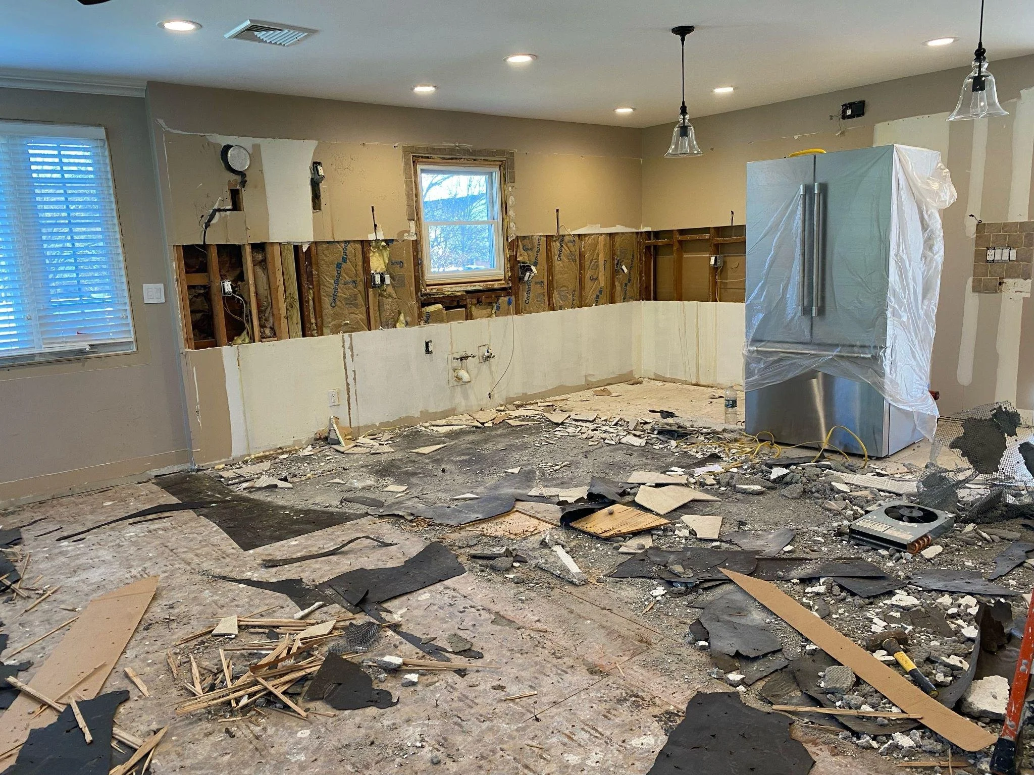 Kitchen undergoing renovation with partially demolished wall, exposed wiring, debris on the floor, and a covered refrigerator.