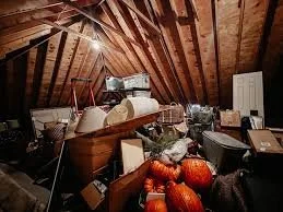 Cluttered attic with various household items and furniture, including pumpkins, a white cabinet, and a wooden bed frame.