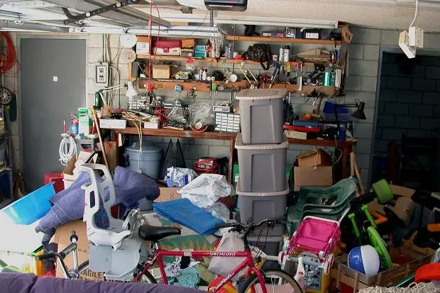 A cluttered garage with shelves holding various tools and containers, stacks of plastic storage bins, a child's pink and green bike, an air compressor, and assorted miscellaneous items.