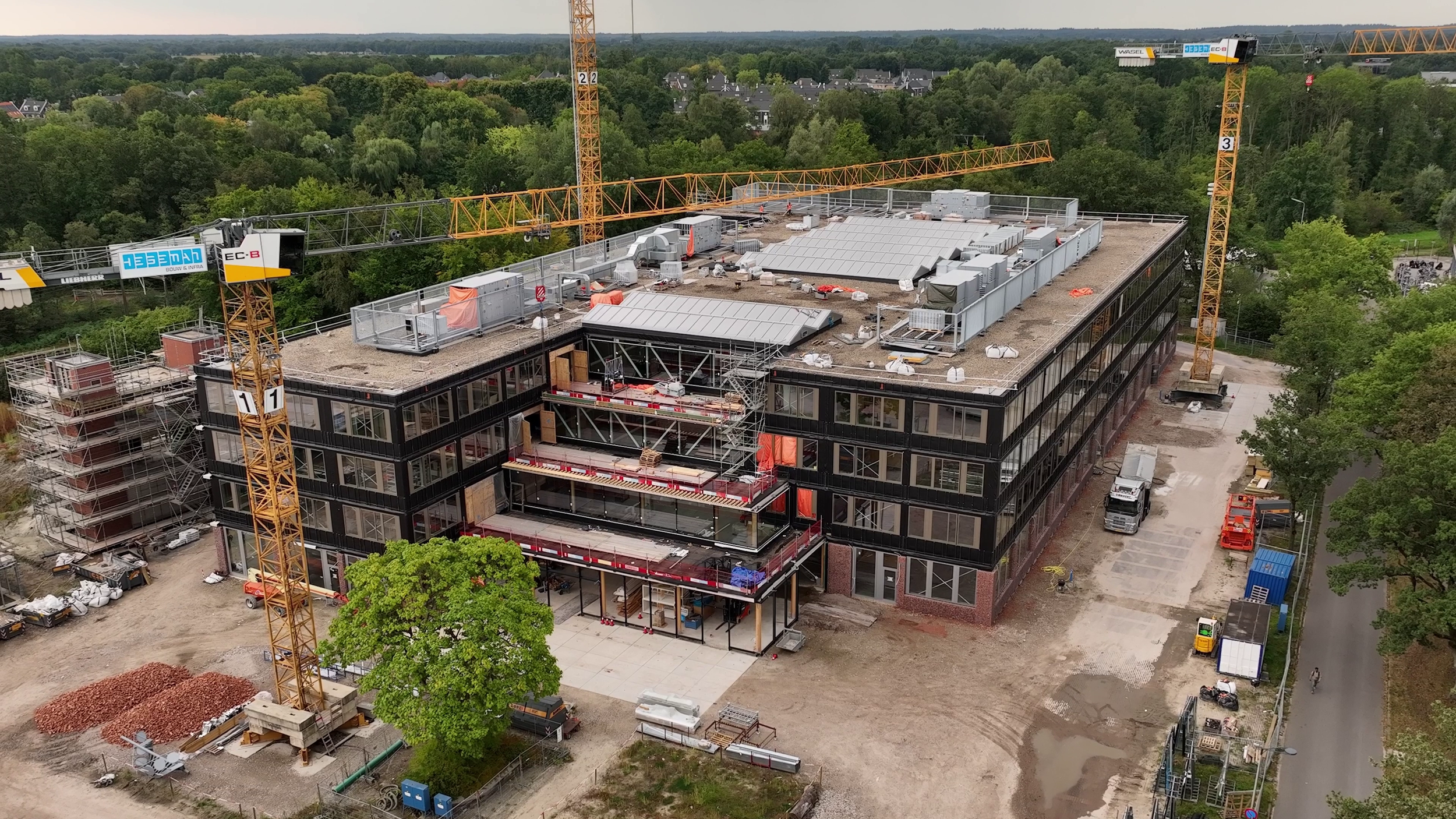 Aerial view of a building under construction with multiple construction cranes and scaffolding, surrounded by trees and neighborhood houses.