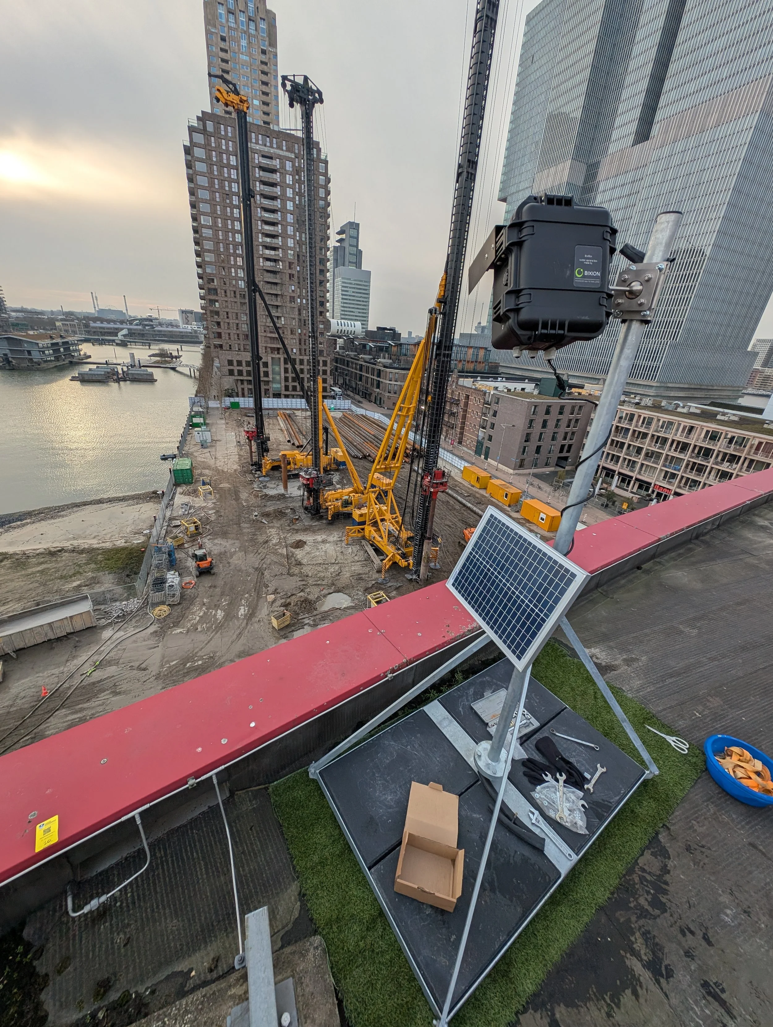 View of a construction site with cranes, construction tools, and a timelapse camera in an urban setting with high-rise buildings, water, and cloudy sky in the background.