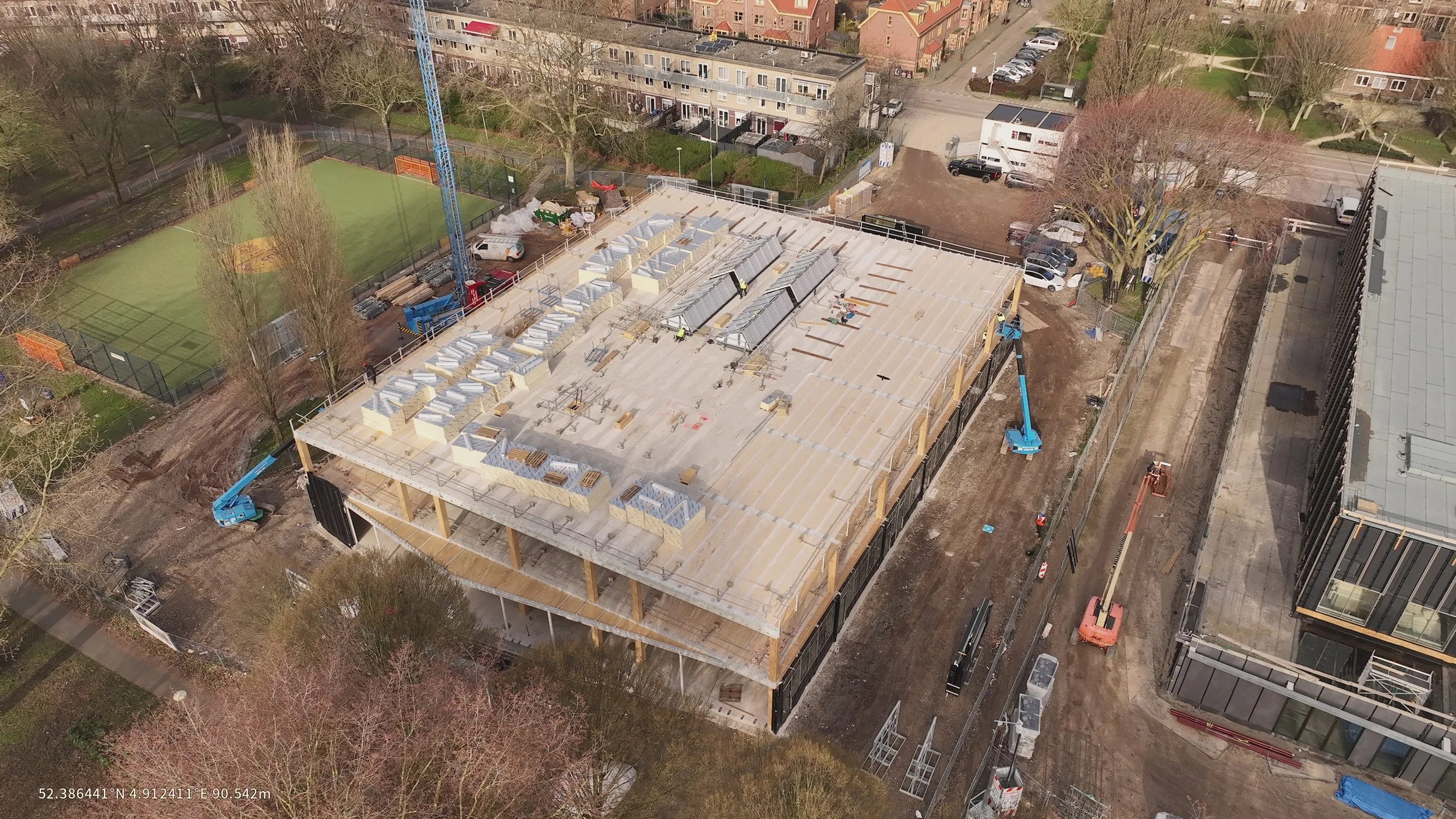 Aerial view of a building under construction with workers, construction materials, crane, and equipment, surrounded by trees, parked cars, and residential buildings.