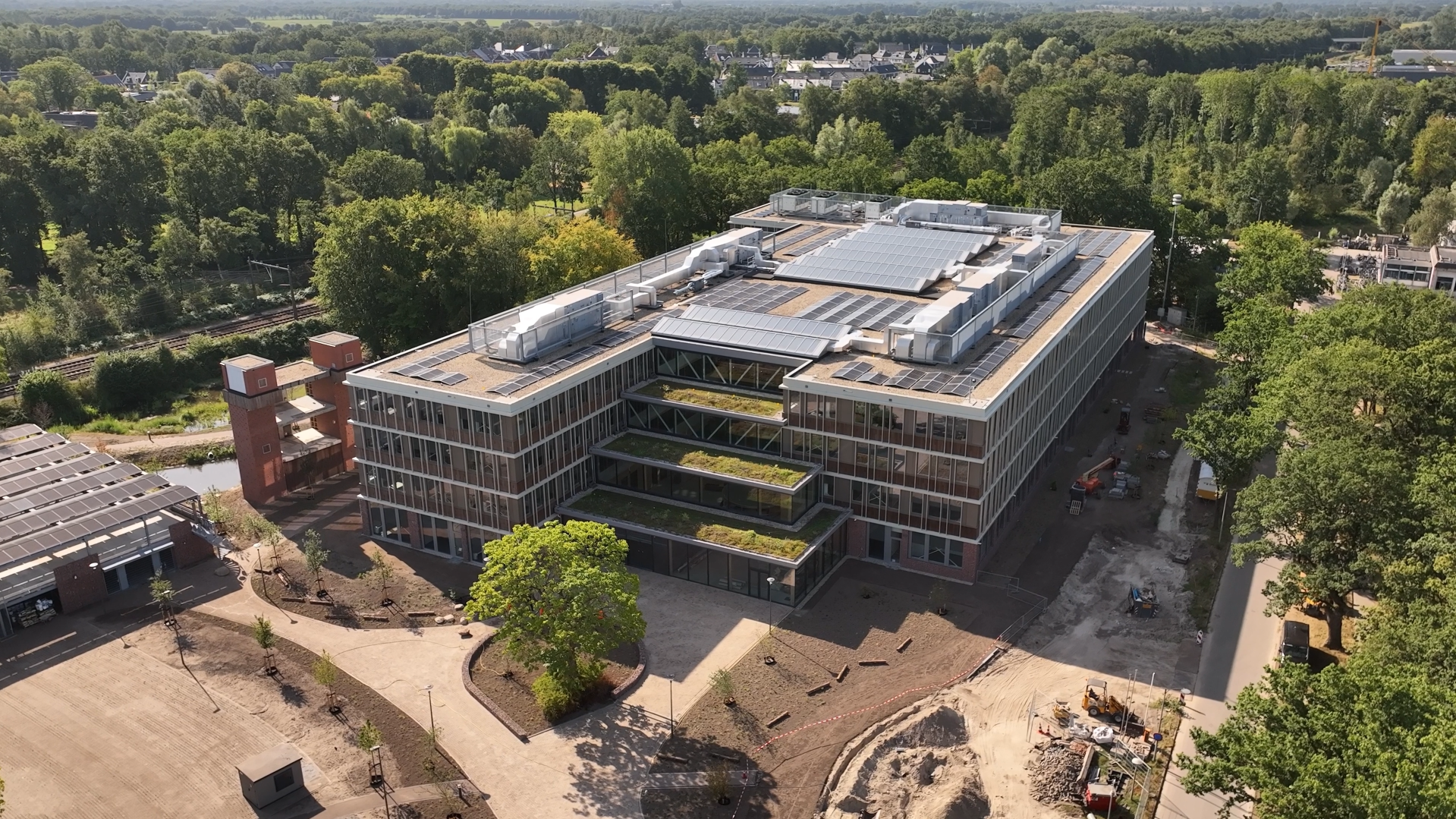 An aerial view of a modern building with solar panels on the roof, surrounded by trees and construction area.