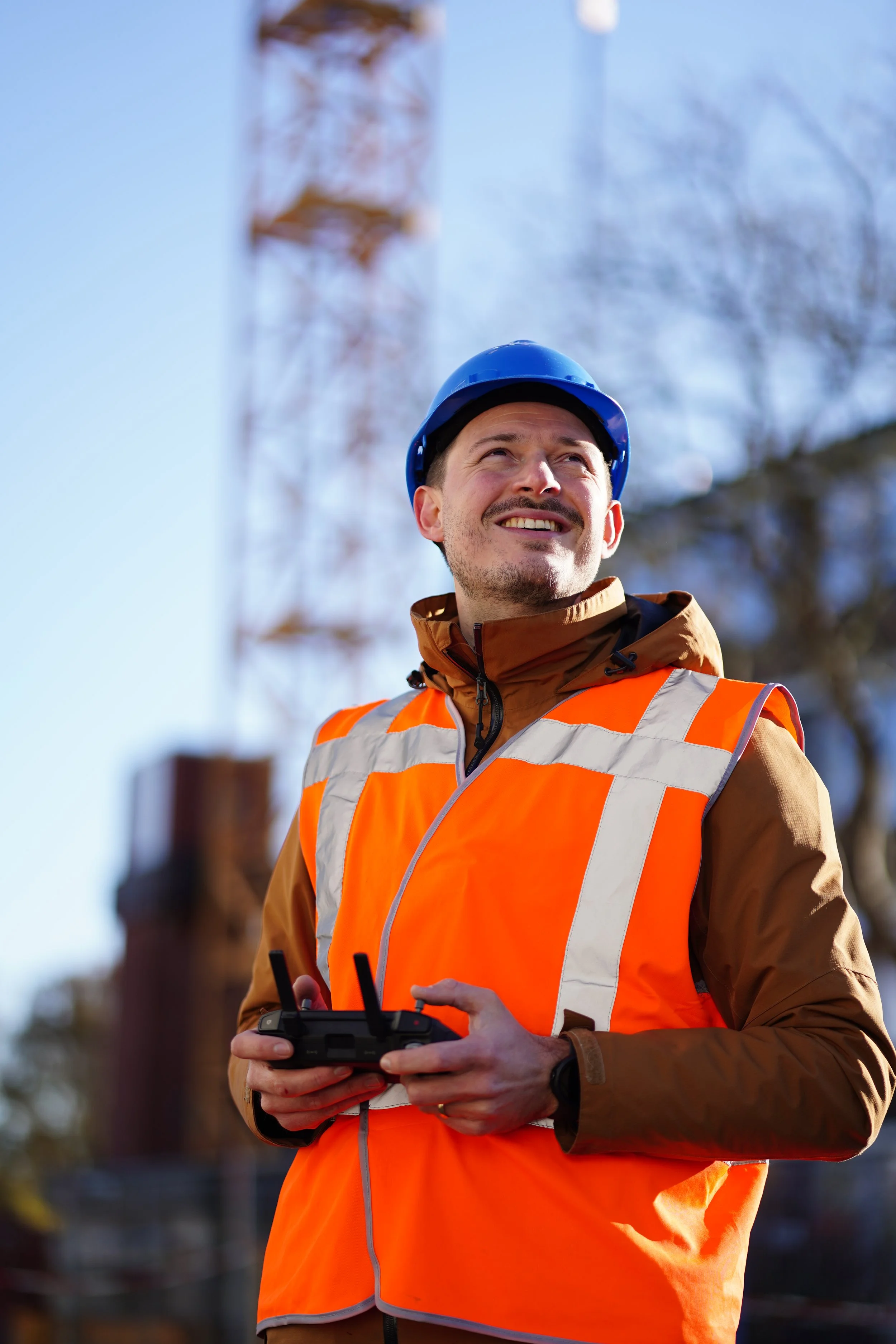 A smiling man wearing a blue safety helmet and an orange safety vest holding a remote control, standing outdoors during daytime.